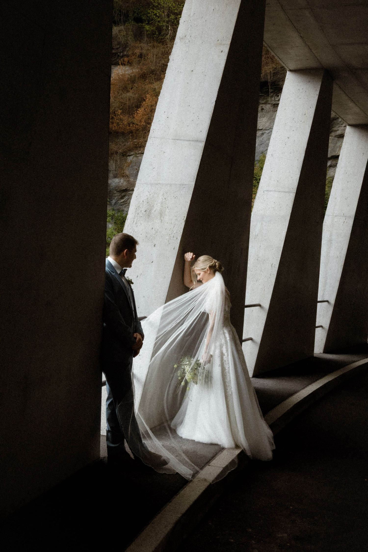Bride and groom share intimate moment between concrete pillars, veil flowing dramatically.
