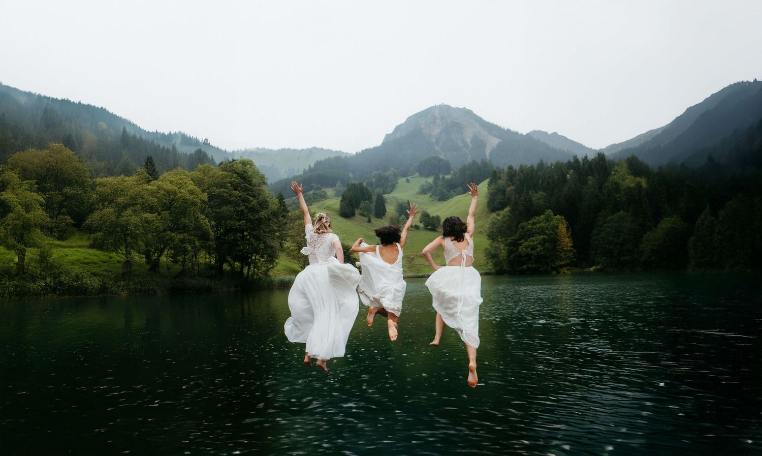 Three women in white dresses joyfully jump into a lake surrounded by mountains.