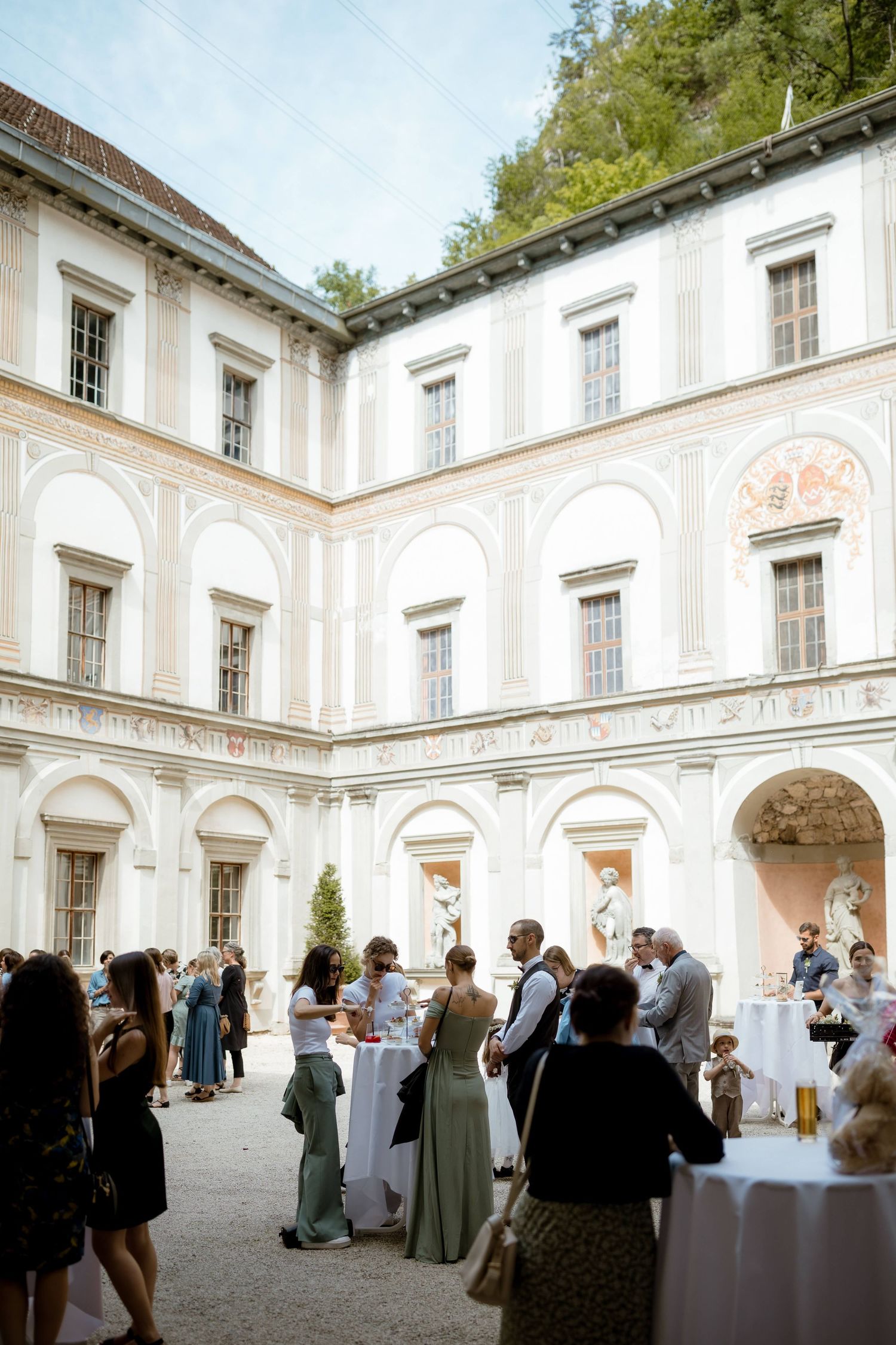 Elegant wedding reception in a classical Italian courtyard with white arched architecture and guests mingling around tables.