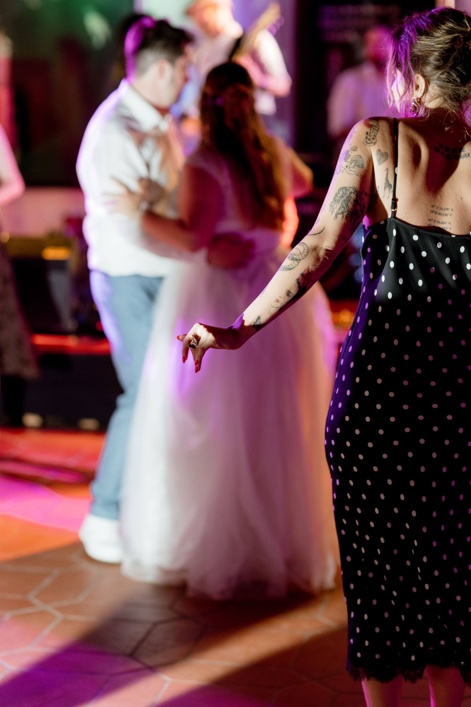 A tattooed woman in a polka dot dress watches the bride and groom share a dance on the wedding floor.
