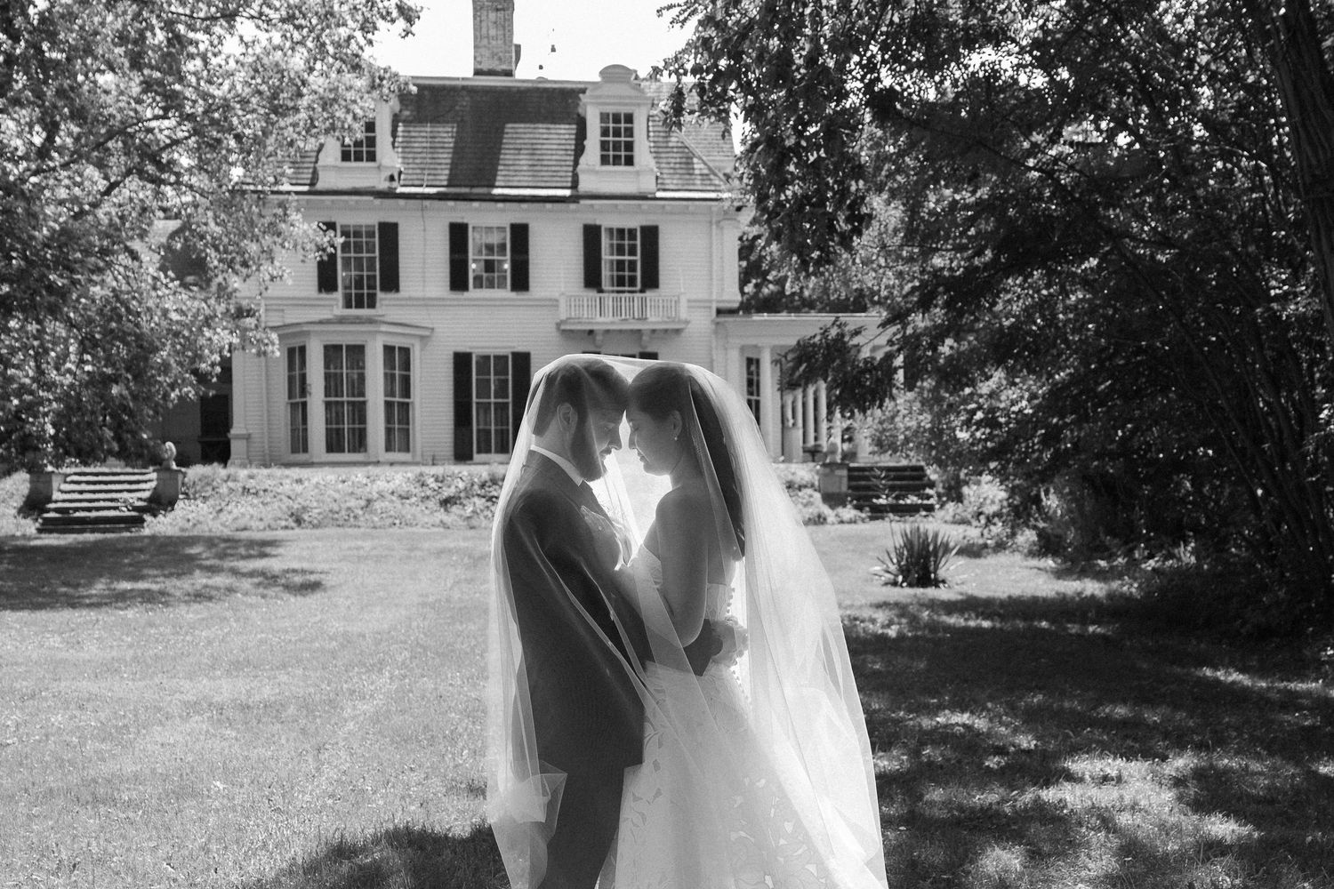 Black and white wide shot of bride and groom standing face-to-face beneath the wedding veil on the lawn with Wadsworth Homestead in the back