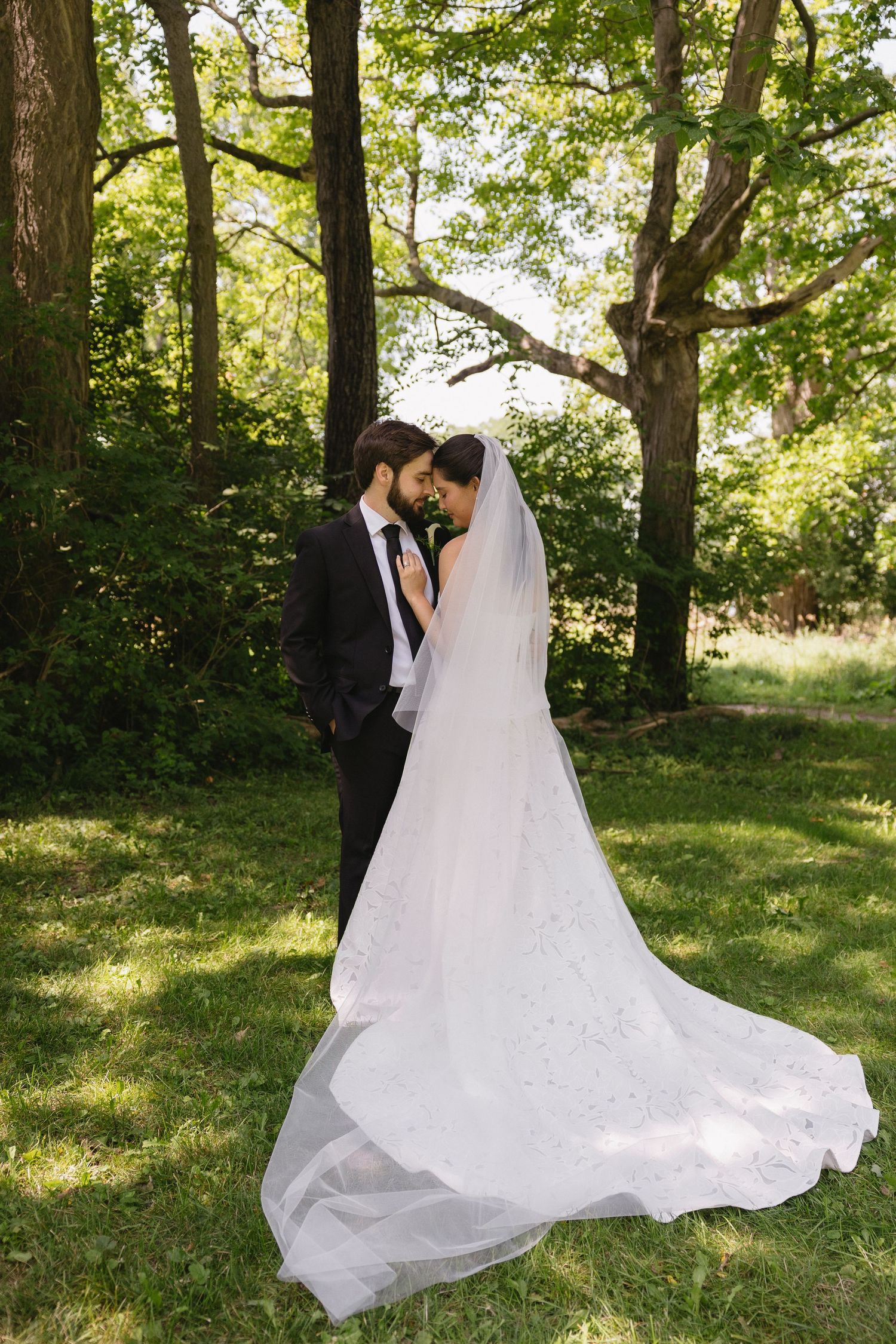 Bride and groom embracing in a wooded area with the bride's long veil spread across the lawn at Wadsworth Homestead in Geneseo, NY.