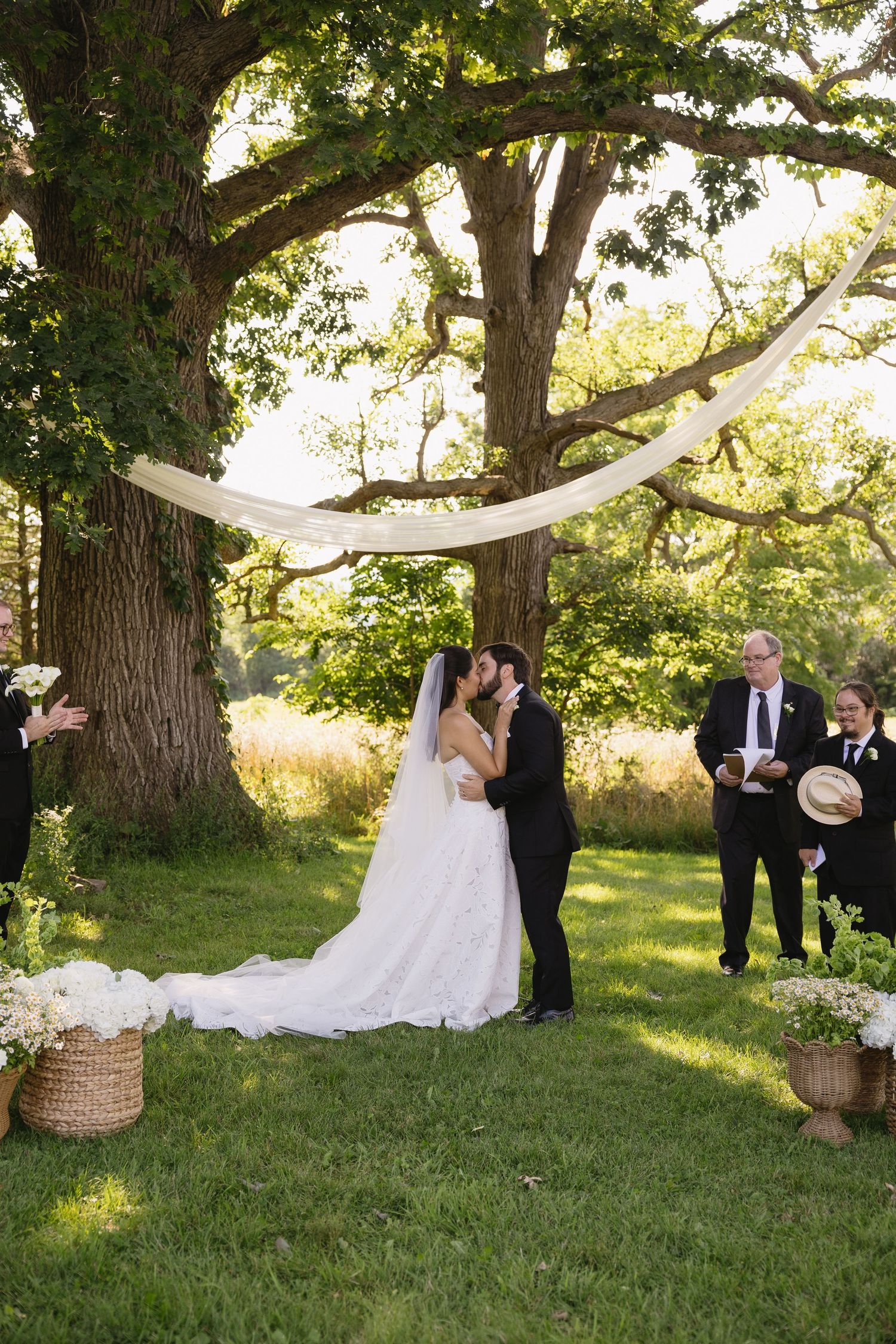 Bride and groom first kiss during outdoor ceremony under oak trees at Wadsworth Homestead in Geneseo, NY.