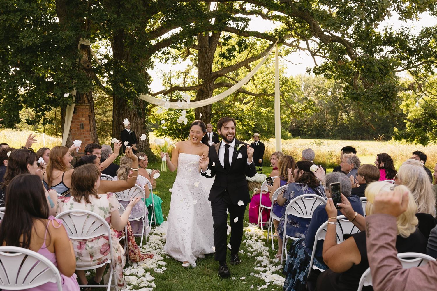 Bride and groom walking up the aisle surrounded by guests tossing white flower petals at outdoor ceremony at Wadsworth Homestead in Geneseo,