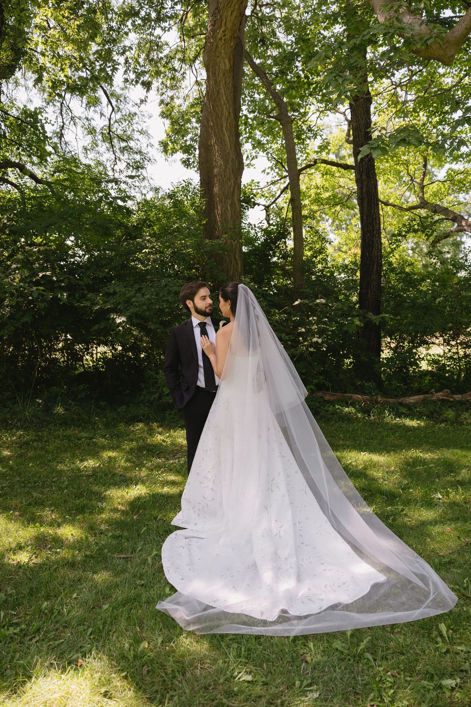 Bride and groom embracing in a wooded area with the bride's long veil spread across the lawn at Wadsworth Homestead in Geneseo, NY.