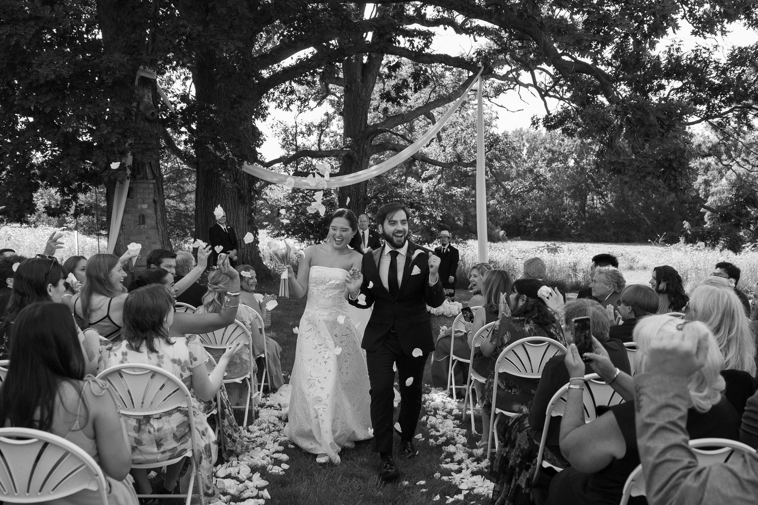 Black and white photo of bride and groom smiling as guests toss flower petals during outdoor ceremony recessional at Wadsworth Homestead in