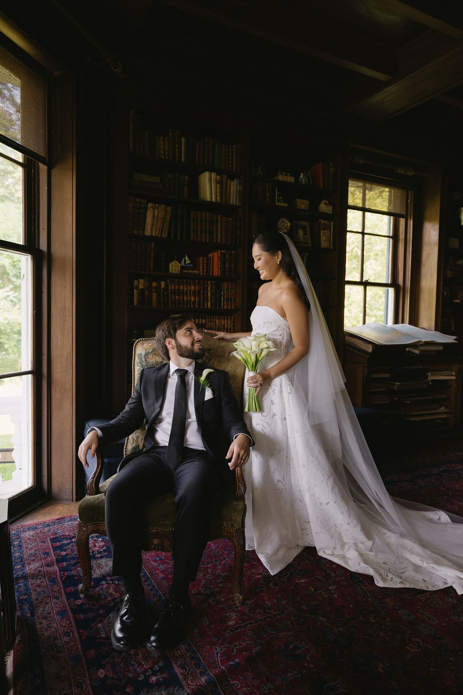 Bride standing with bouquet next to groom seated in armchair inside historic library at Wadsworth Homestead in Geneseo, NY.