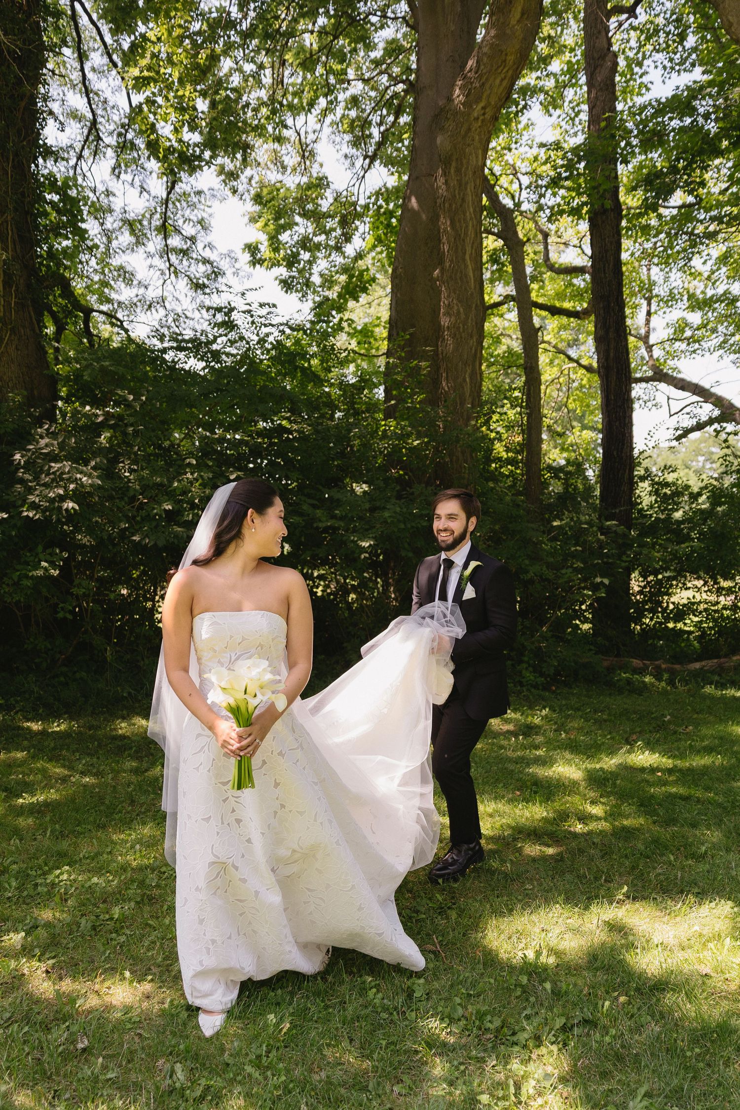 Candid moment of groom holding bride's dress train as they walk through a wooded area at Wadsworth Homestead in Geneseo, NY.