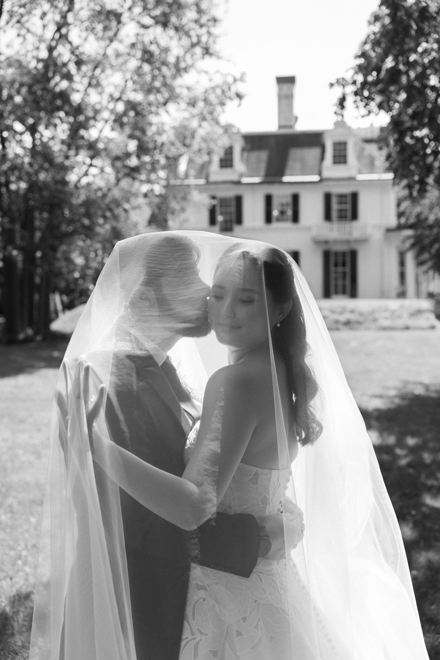 Black and white shot of groom kissing bride on the cheek beneath her wedding veil with Wadsworth Homestead in the background in Geneseo, NY.