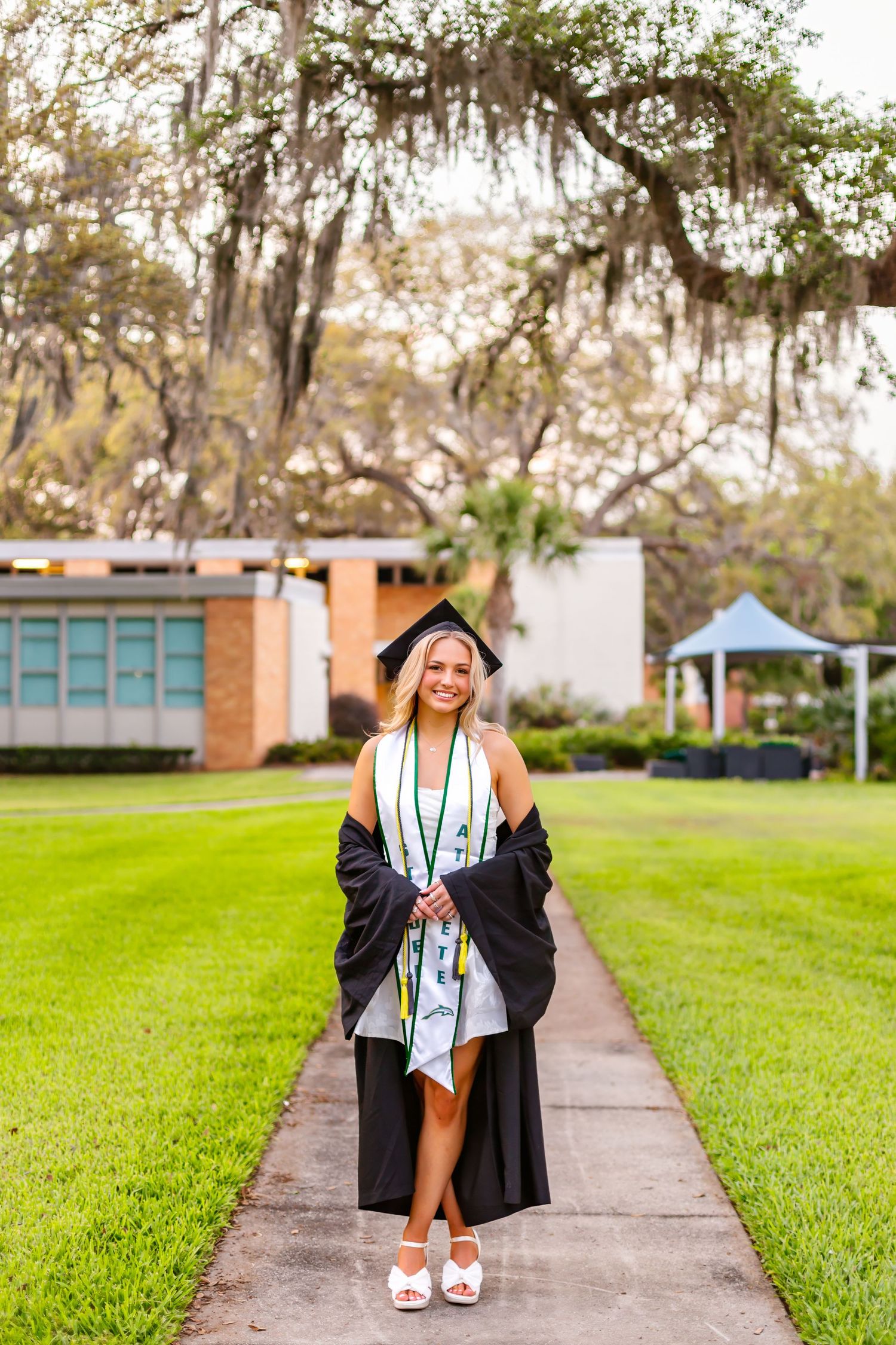 Smiling blonde graduate in black cap and gown walking on campus pathway beneath moss-draped oak trees.
