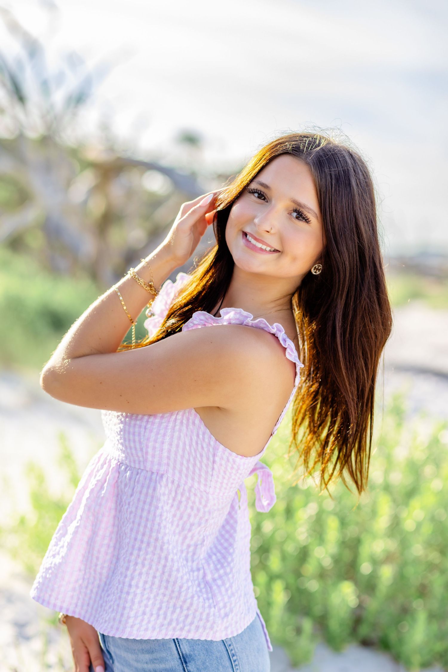Smiling brunette teen girl in pink top posing outdoors near beach vegetation.