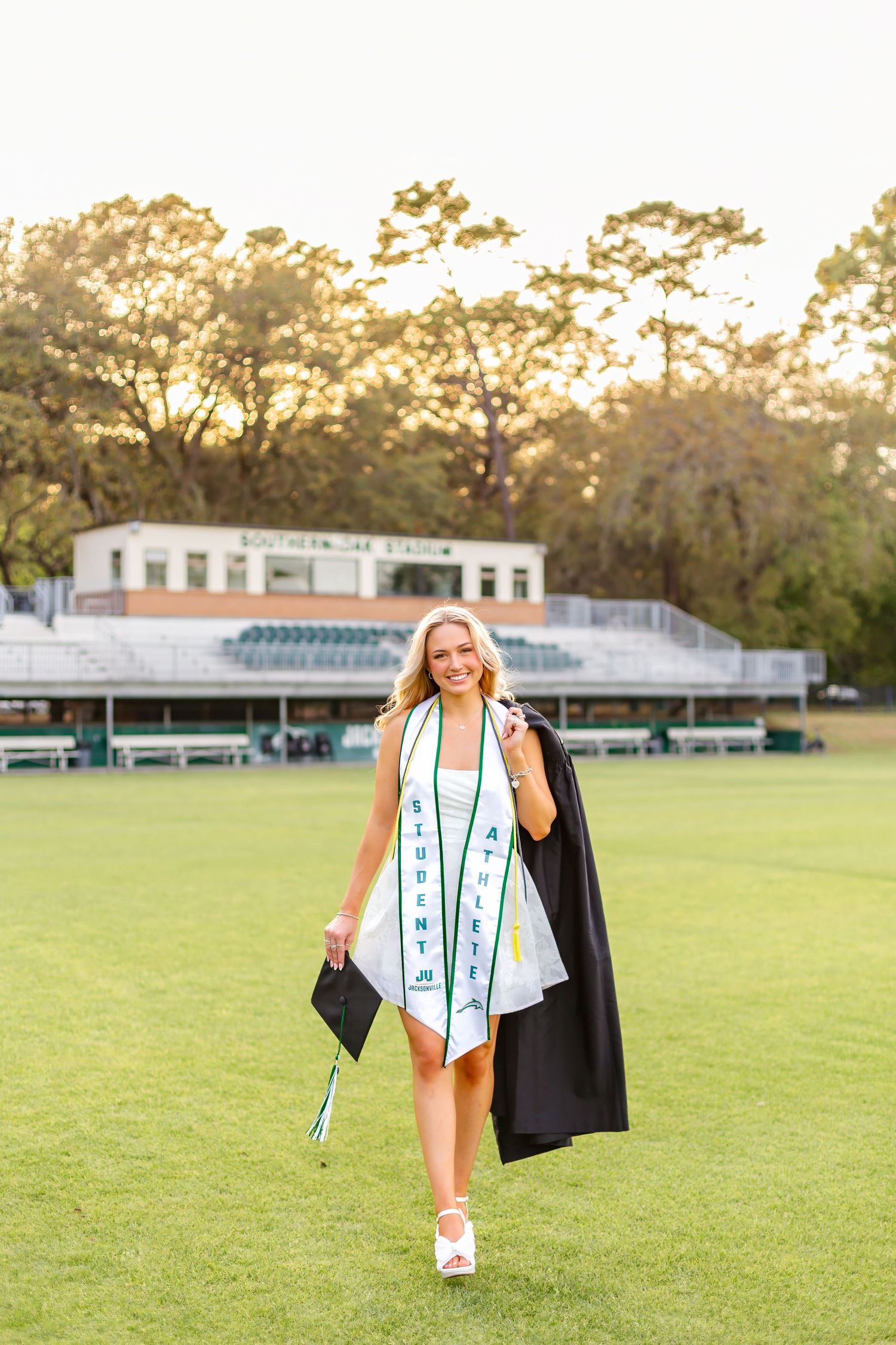 Smiling blonde female graduate in cap and gown walking across green stadium field at golden hour.