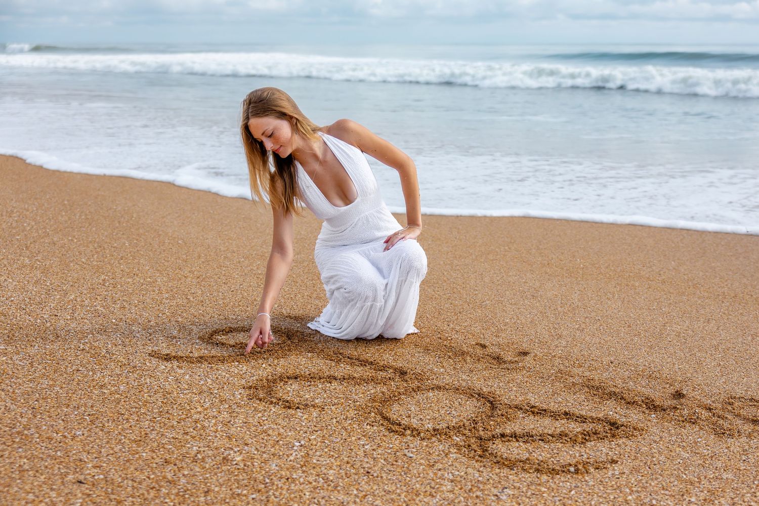 Young woman in white dress writing in sand on sunny beach near waves.