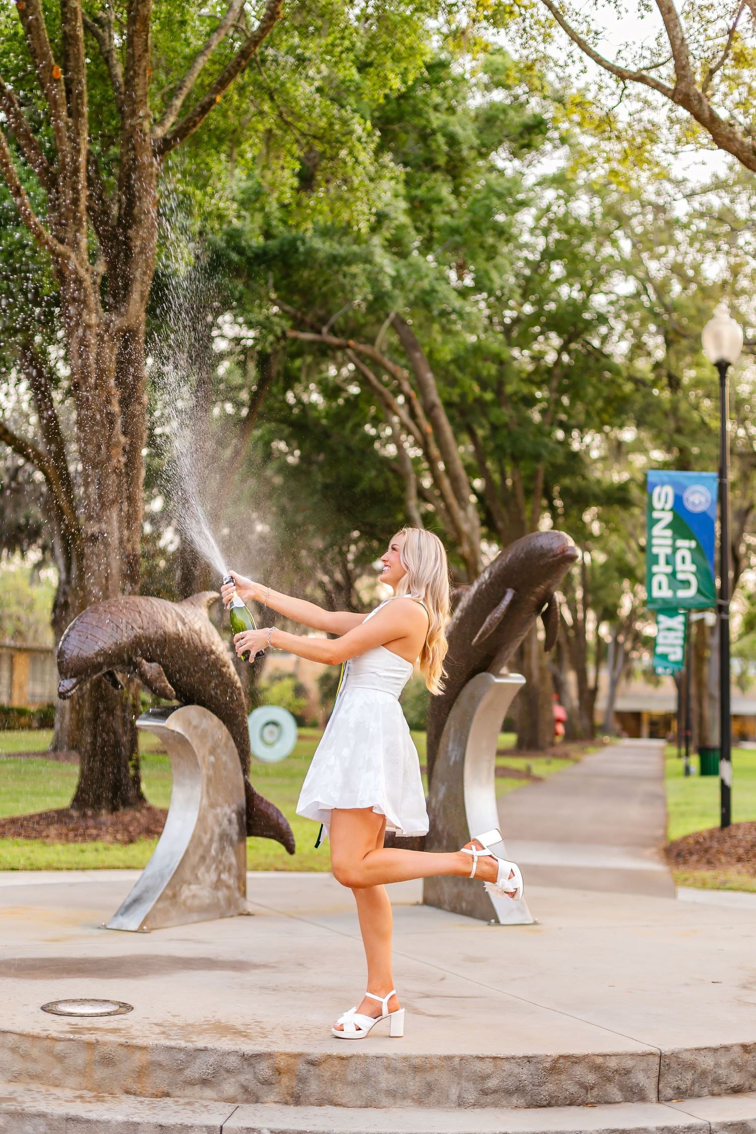 Excited blonde woman in white dress popping champagne near bronze dolphin sculpture in sunny park.