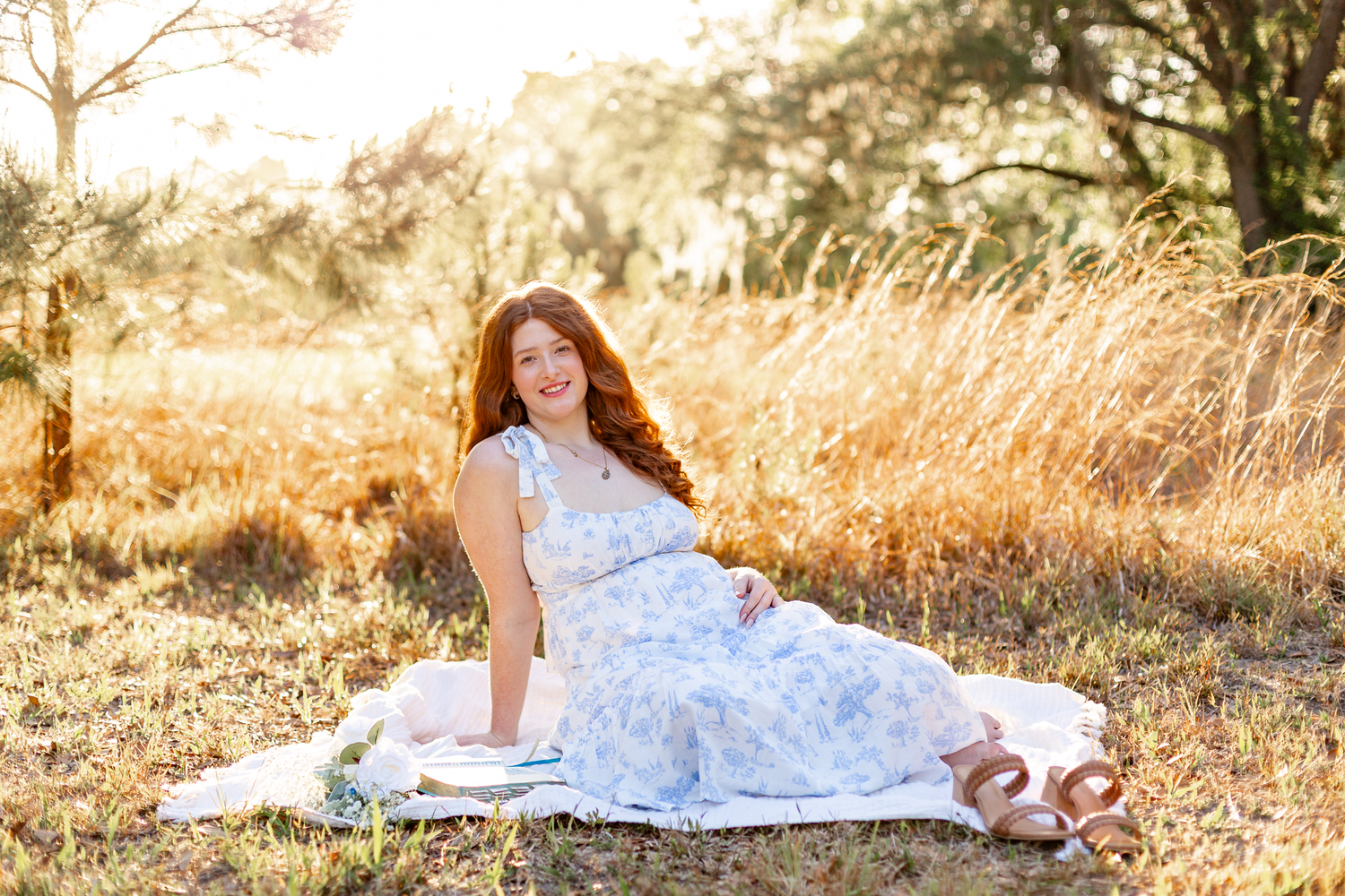 Smiling redhead woman in blue floral dress sits on blanket in golden sunlit field.