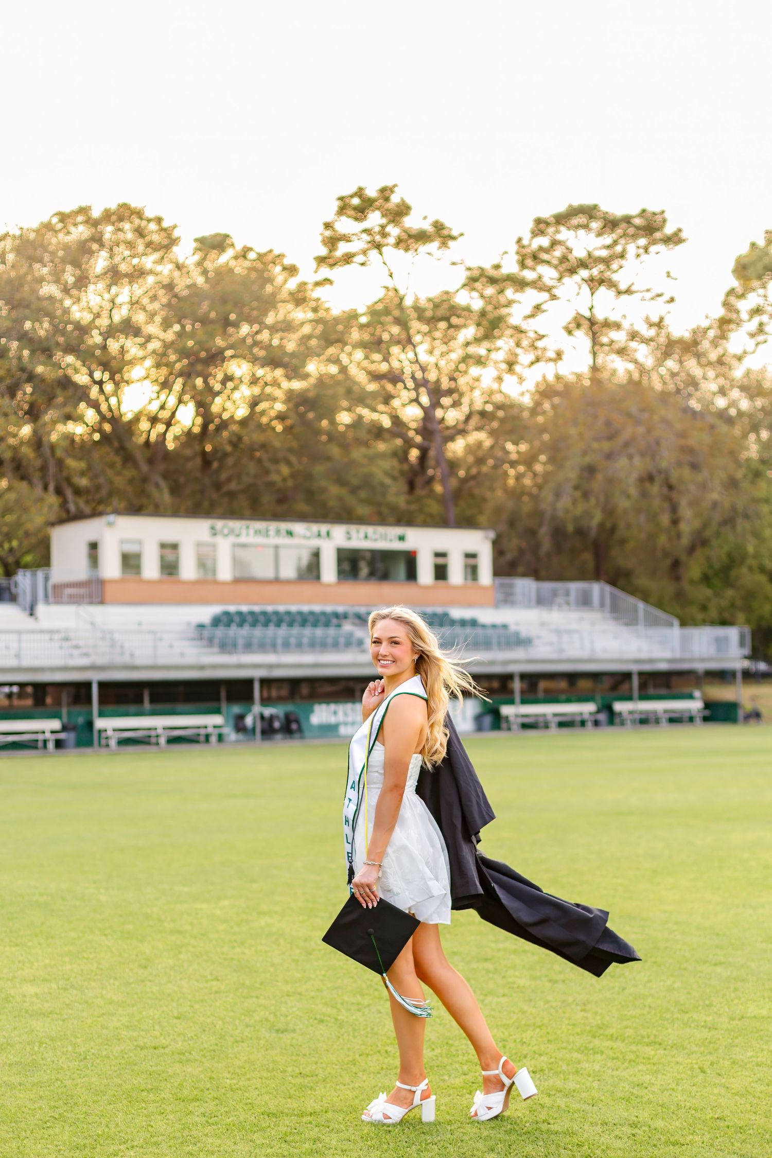 Smiling blonde graduate in white dress carrying cap and gown on stadium field.