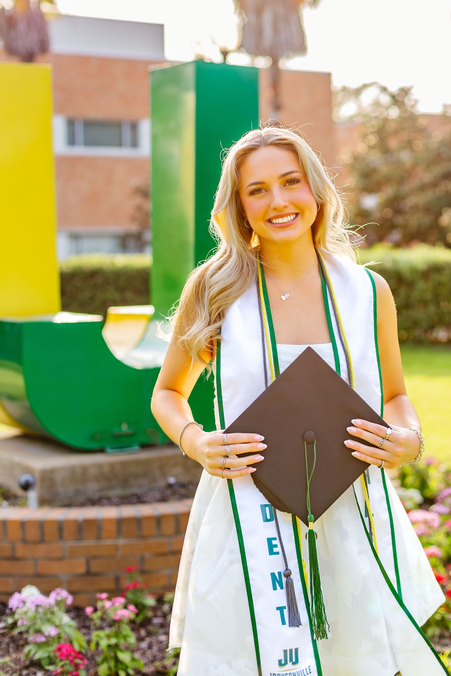 Smiling blonde graduate in white dress holds black mortarboard cap in front of JU campus sculpture.