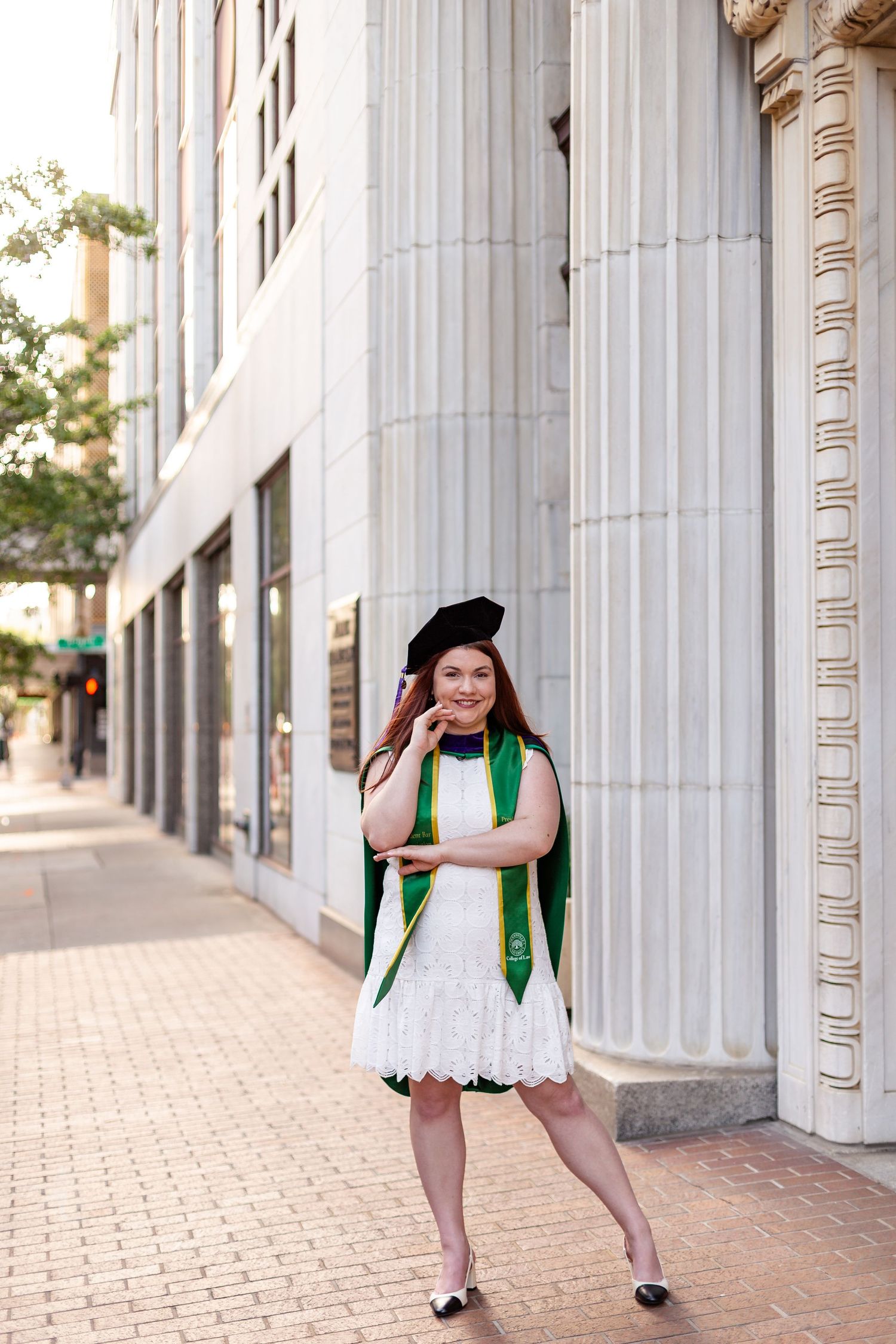 Female law school graduate in white dress, green academic stole, and beret poses on urban sidewalk.