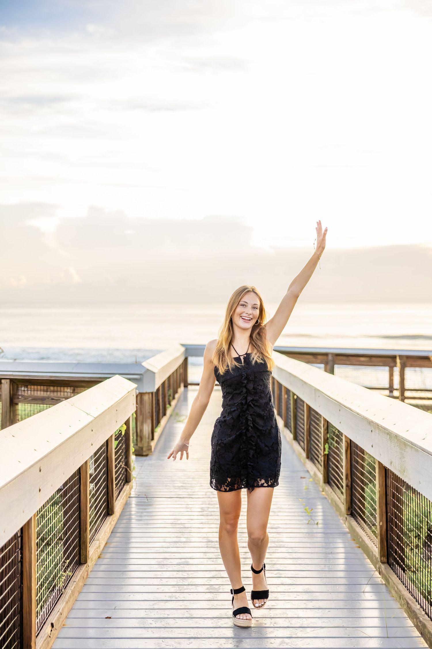 Blonde teen girl in black dress raising arm on wooden boardwalk at sunset.