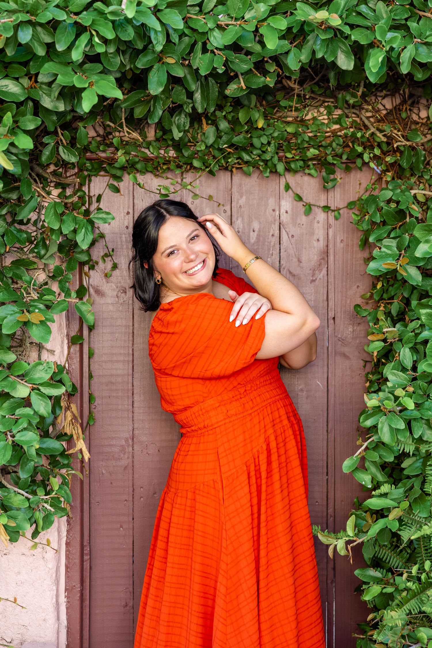 Happy brunette woman in vibrant orange dress posing against rustic wooden door framed by green ivy.