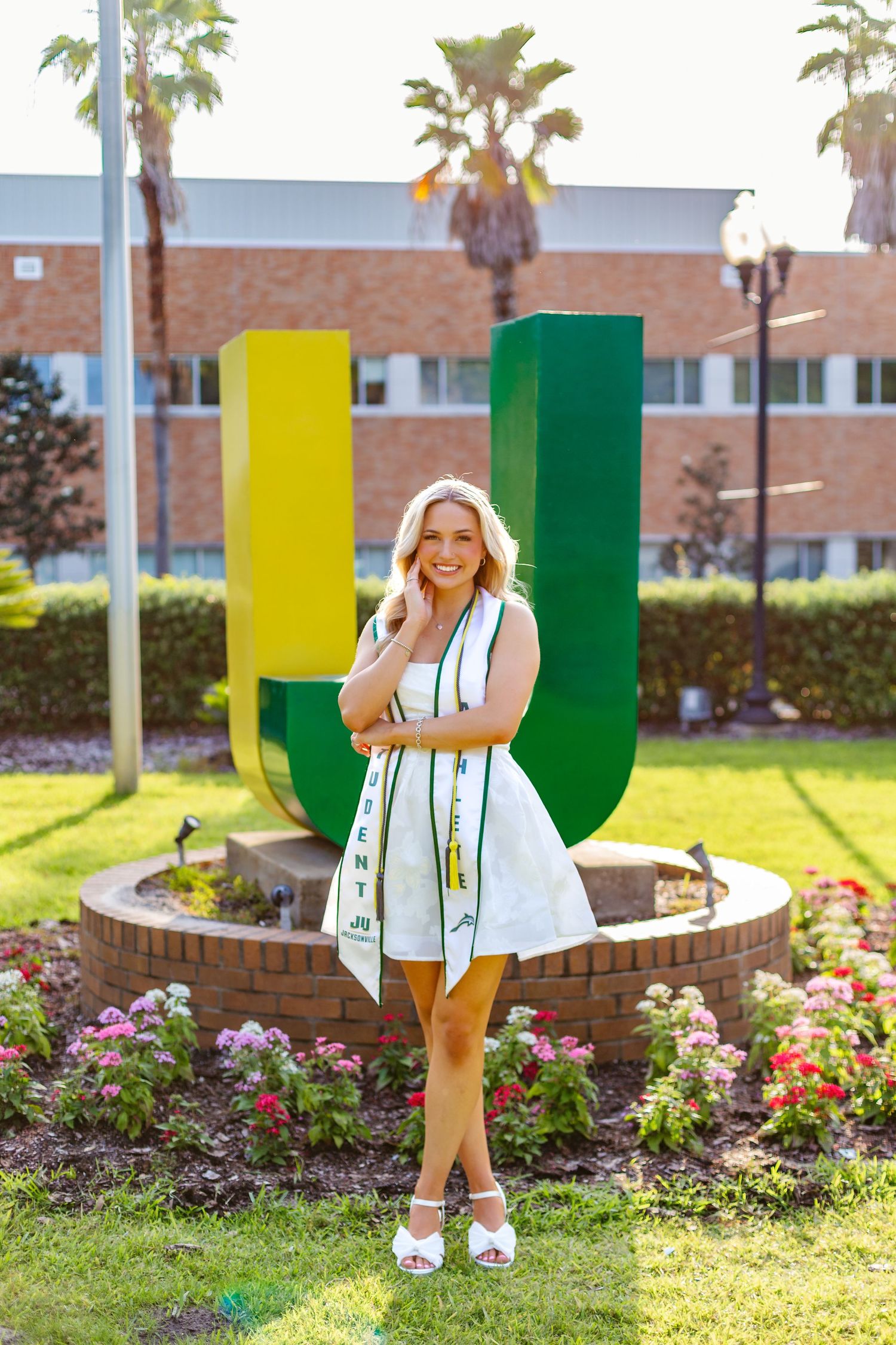 Blonde graduate in white dress and JU sash stands before large green and yellow university letters.