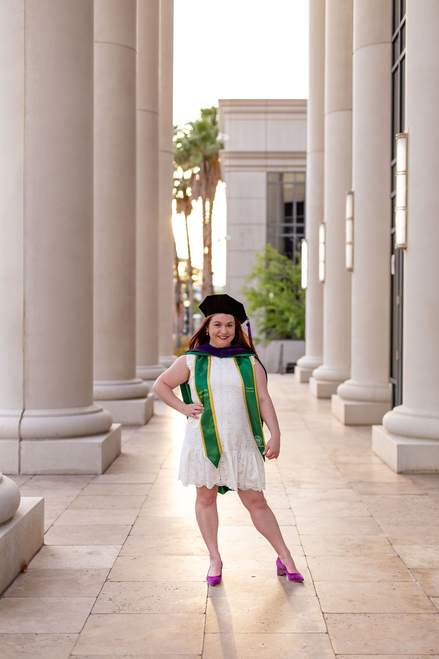 Female law graduate in white dress and green stole poses confidently between tall marble columns.