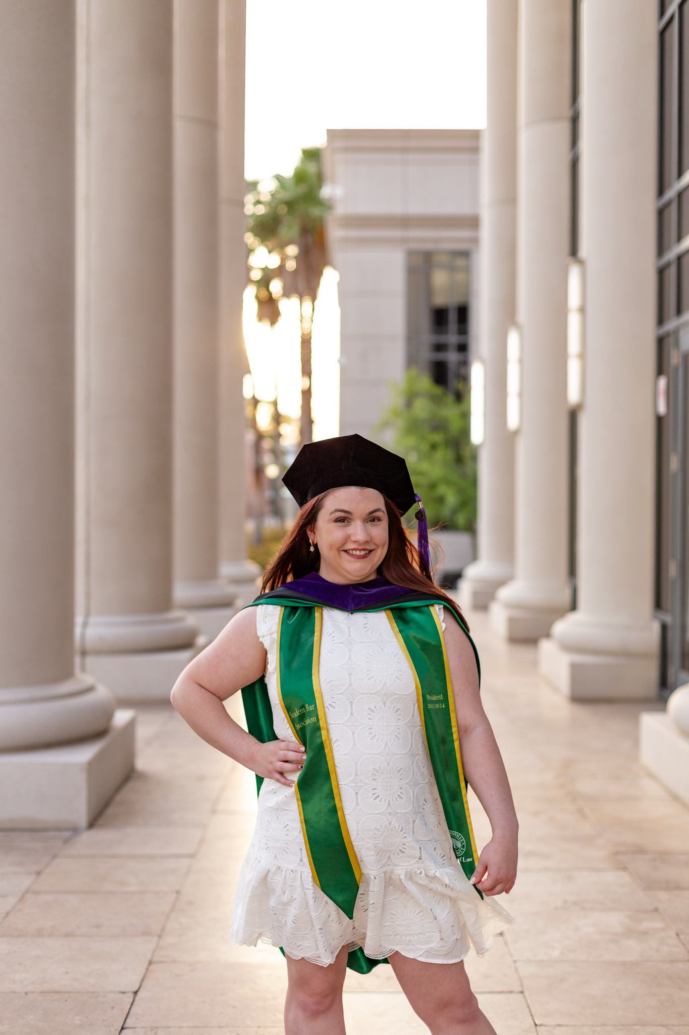 Woman in graduation cap and green doctoral stole posing between columns at sunset.