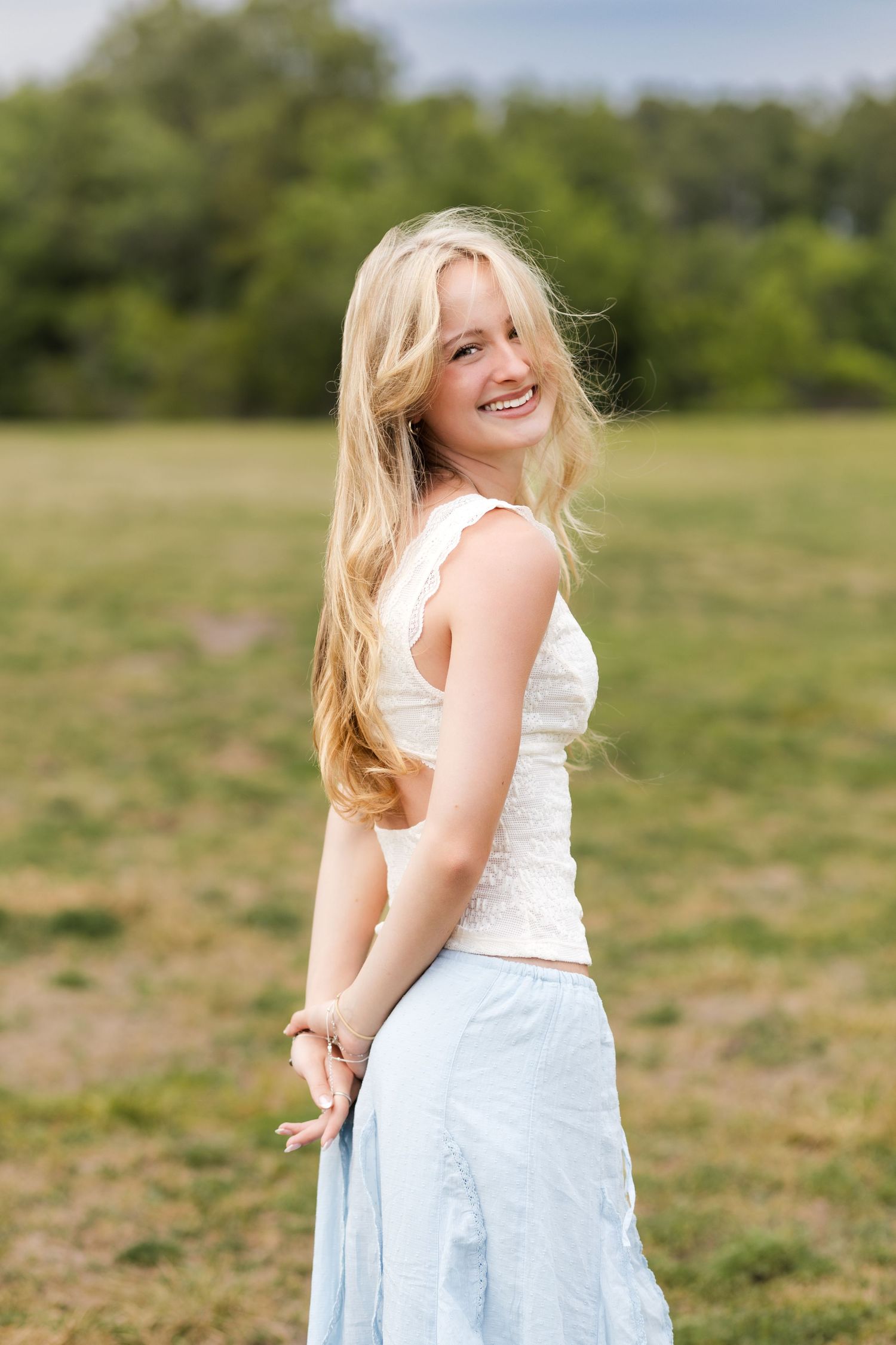 Smiling blonde teen girl in white top and blue skirt posing in a sunny green outdoor field.