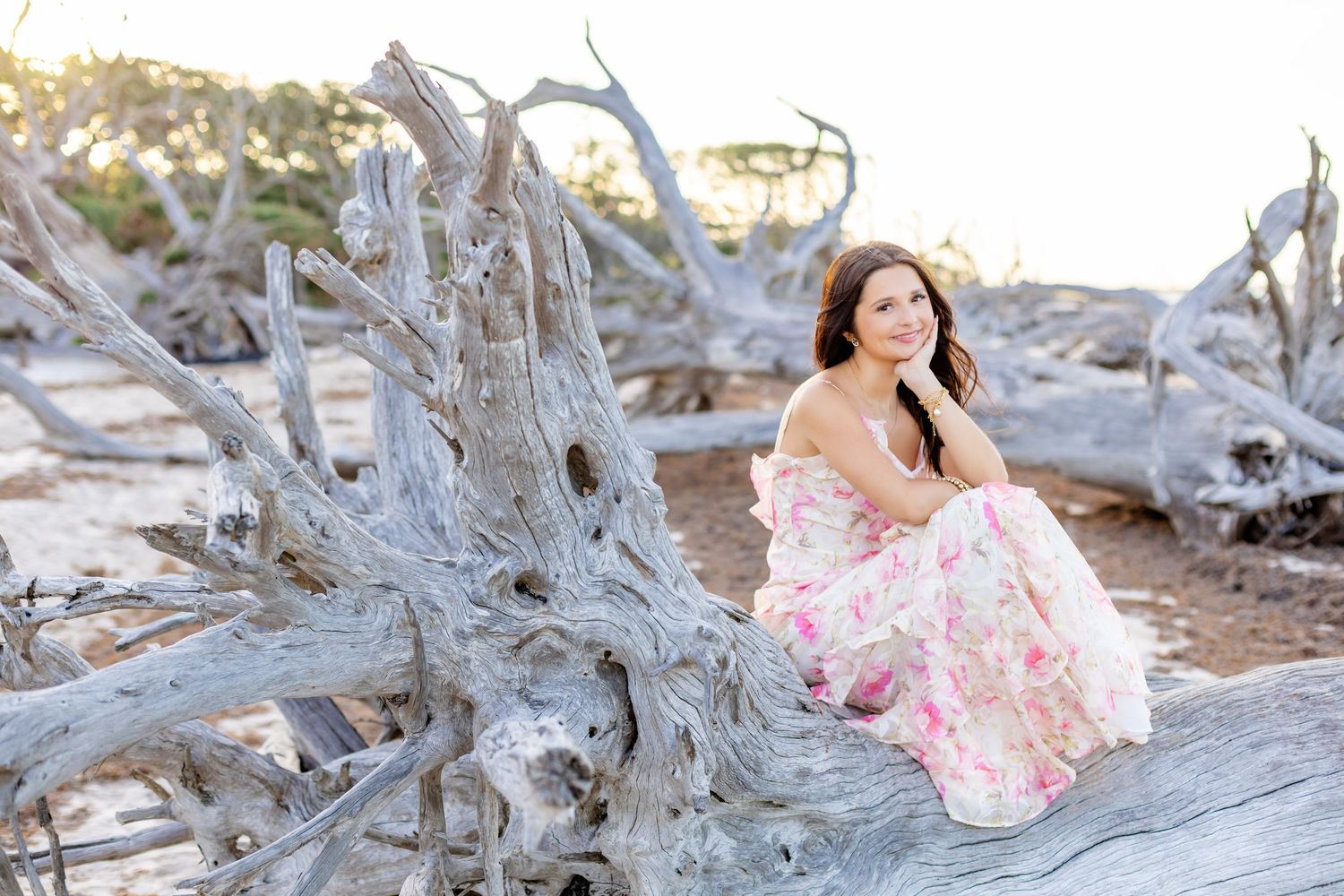 Woman in pink floral gown sitting gracefully on driftwood at a serene beach with pale sand.