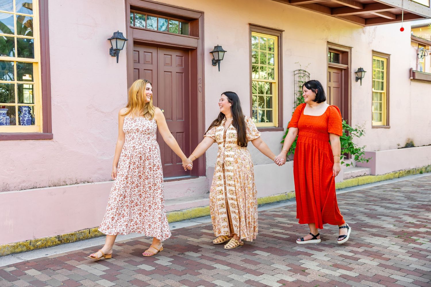 Three women in floral and orange dresses holding hands walking on a cobblestone street outdoors.