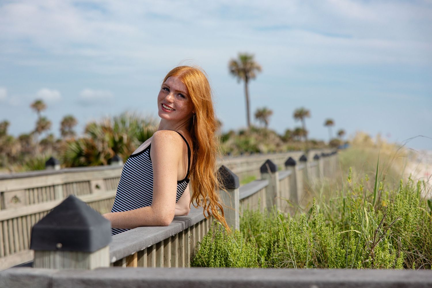 A smiling redhead woman leans on a wooden railing near a beach with palm trees in the background.