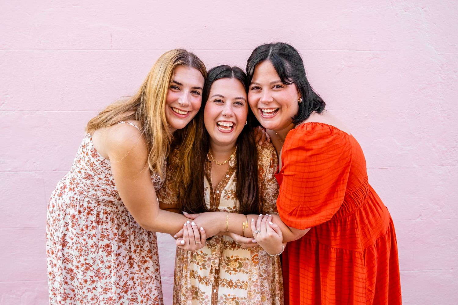 Three smiling young women in colorful dresses hugging together against a pink wall.
