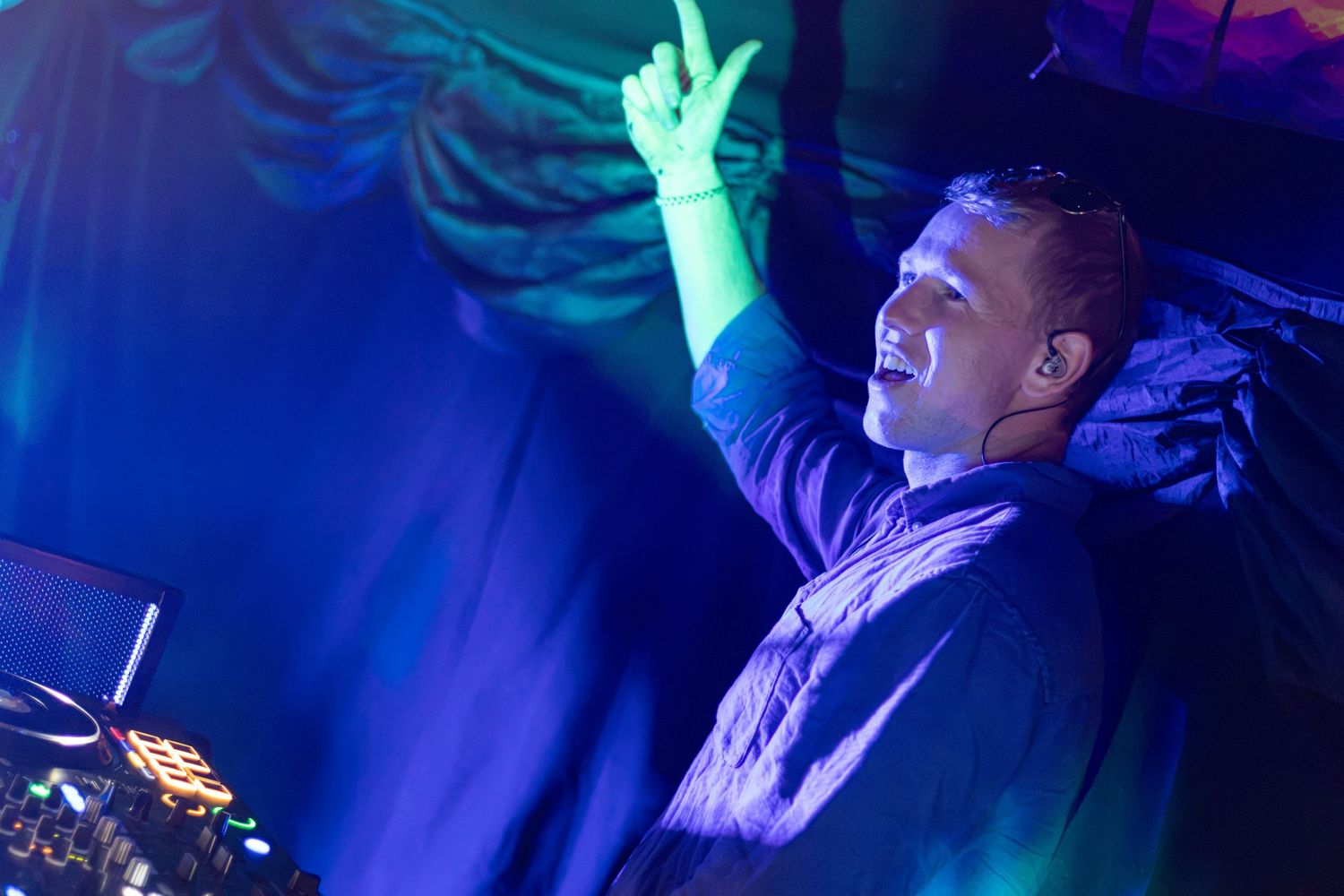 A DJ raises his hand while working at his decks under vibrant blue and green stage lighting at a nightclub.