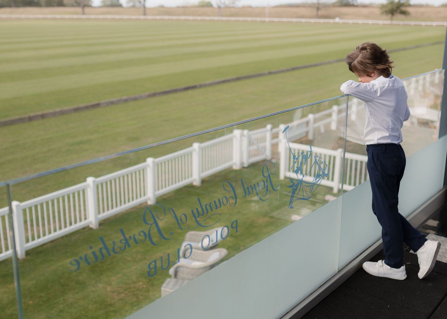 Child standing on balcony overlooking green racecourse with white fencing, peering over glass barrier.