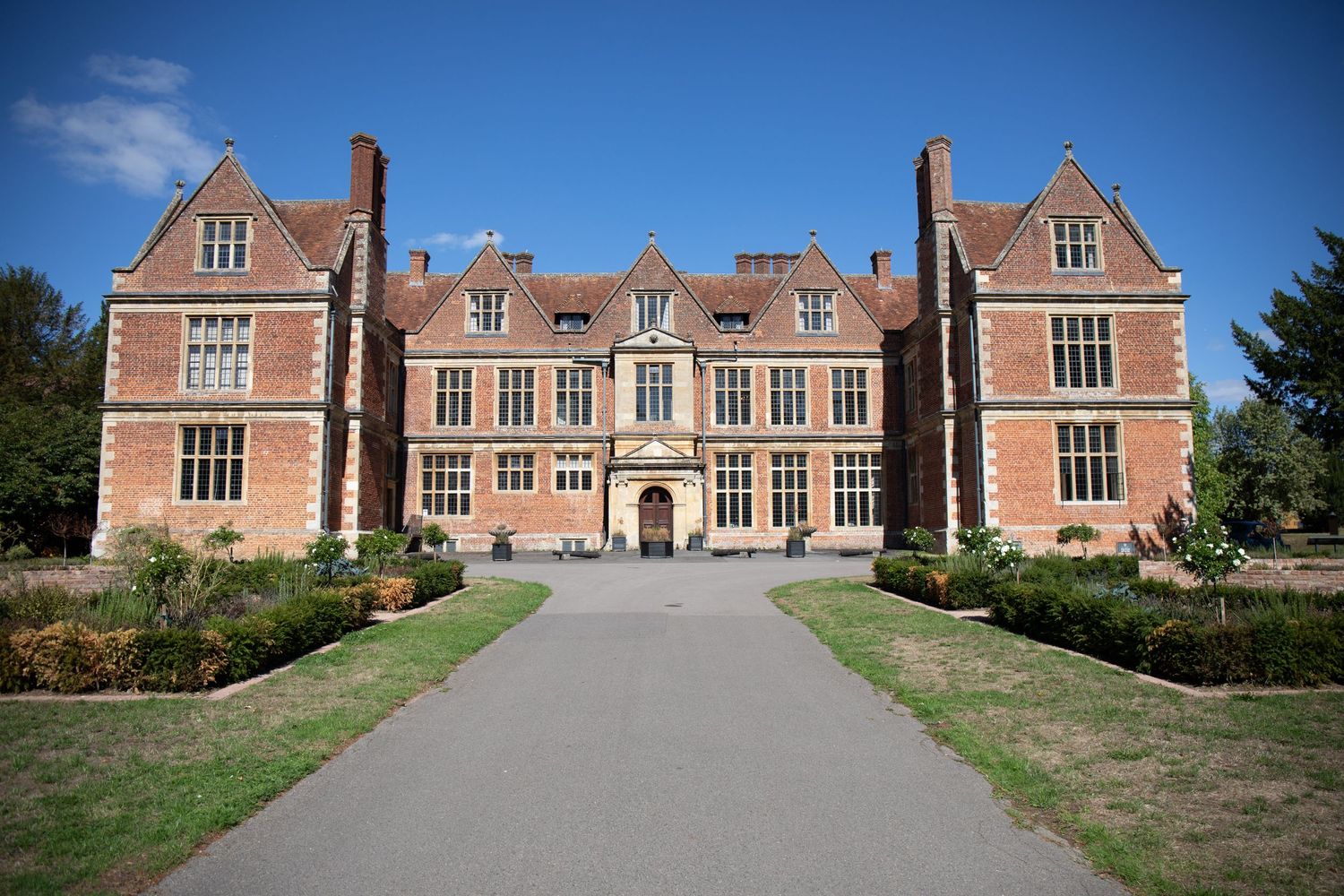 Historic red brick Tudor manor house with symmetrical facade, large windows, and manicured gardens under a clear blue sky.