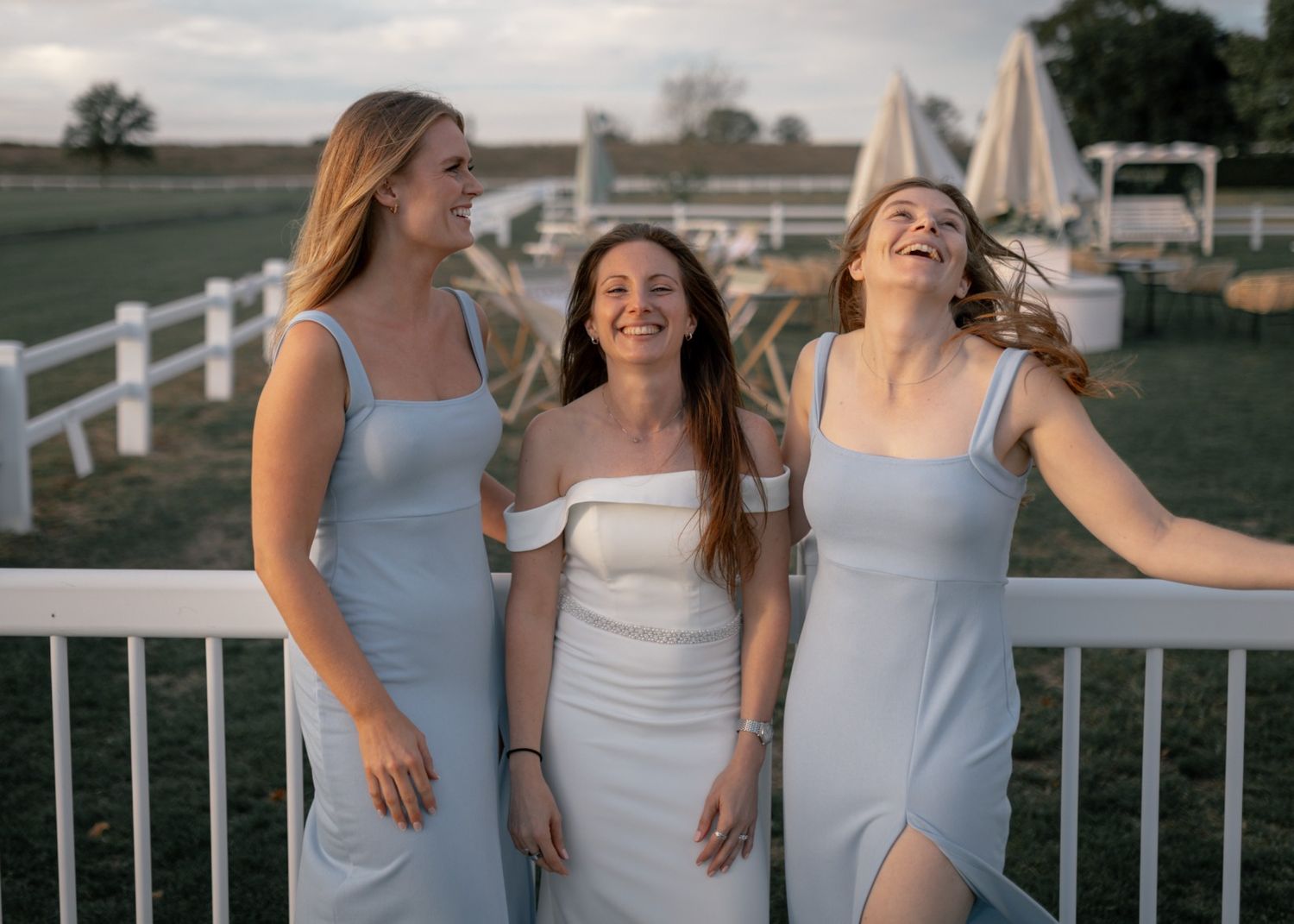 Three young women in light blue and white dresses laughing together outdoors at a wedding venue.