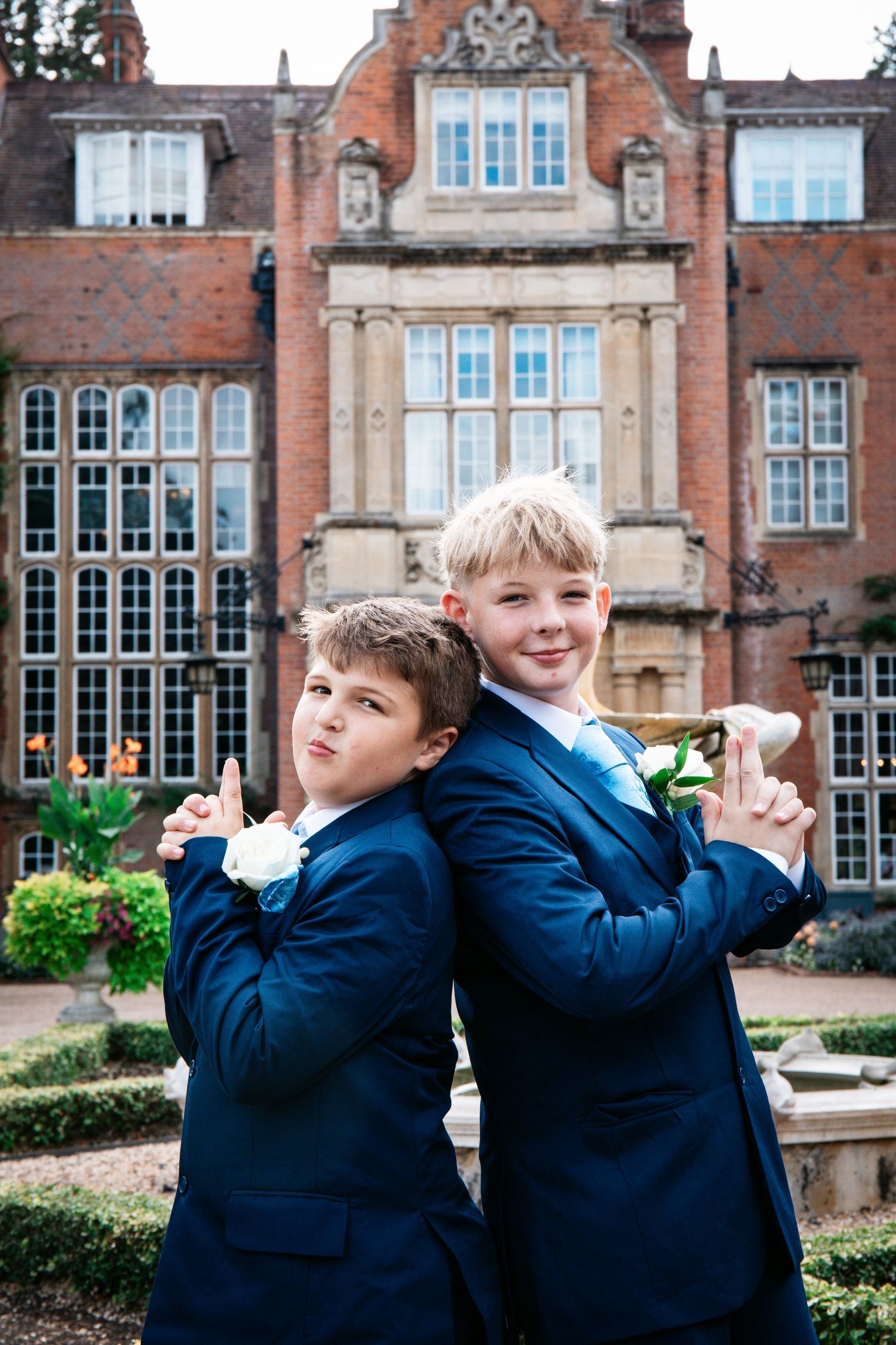 Two boys in navy suits with white boutonnieres pose back-to-back at a wedding outside a grand brick manor.