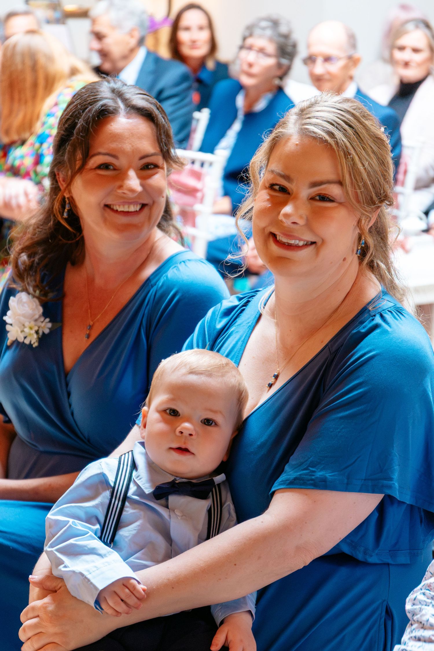 Two bridesmaids in teal dresses smile while holding a baby boy in a bow tie at a wedding ceremony.