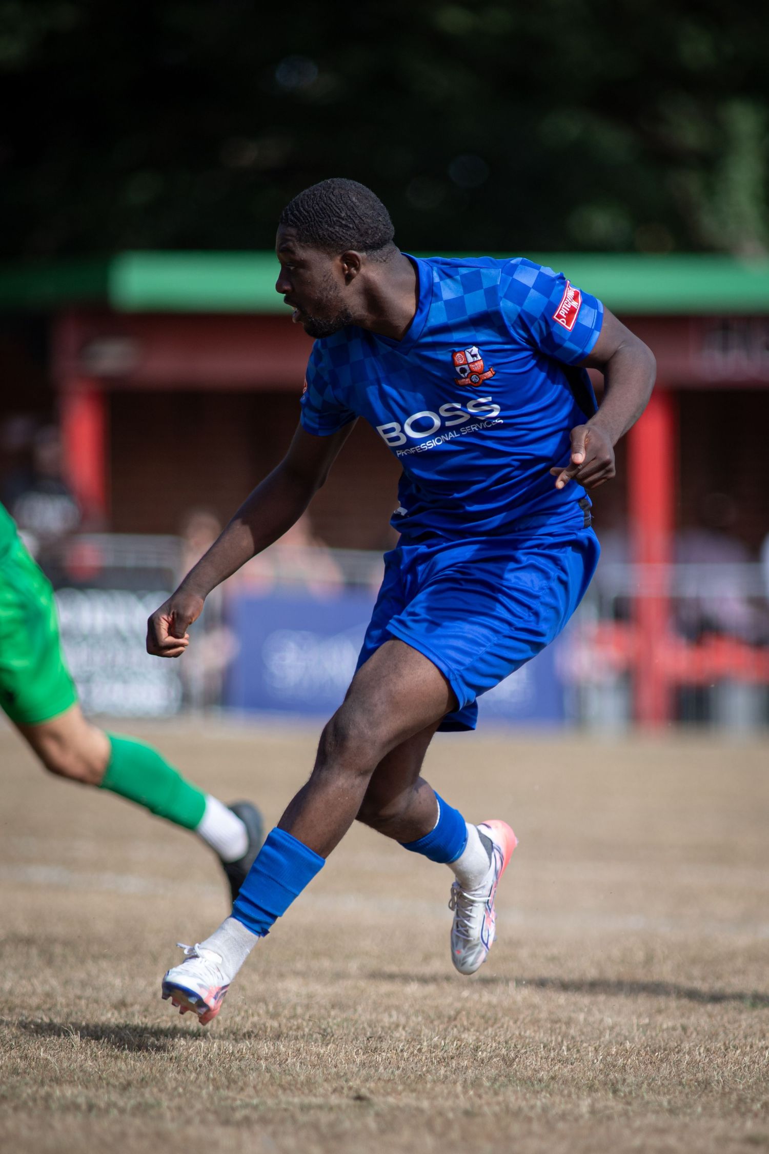 A young footballer in a blue BOSS sponsored kit sprints during a sunny outdoor soccer match.
