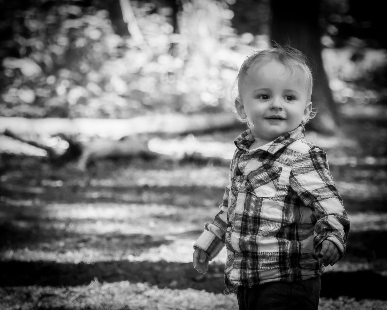 Black and white portrait of a smiling toddler boy in a plaid shirt standing outdoors.