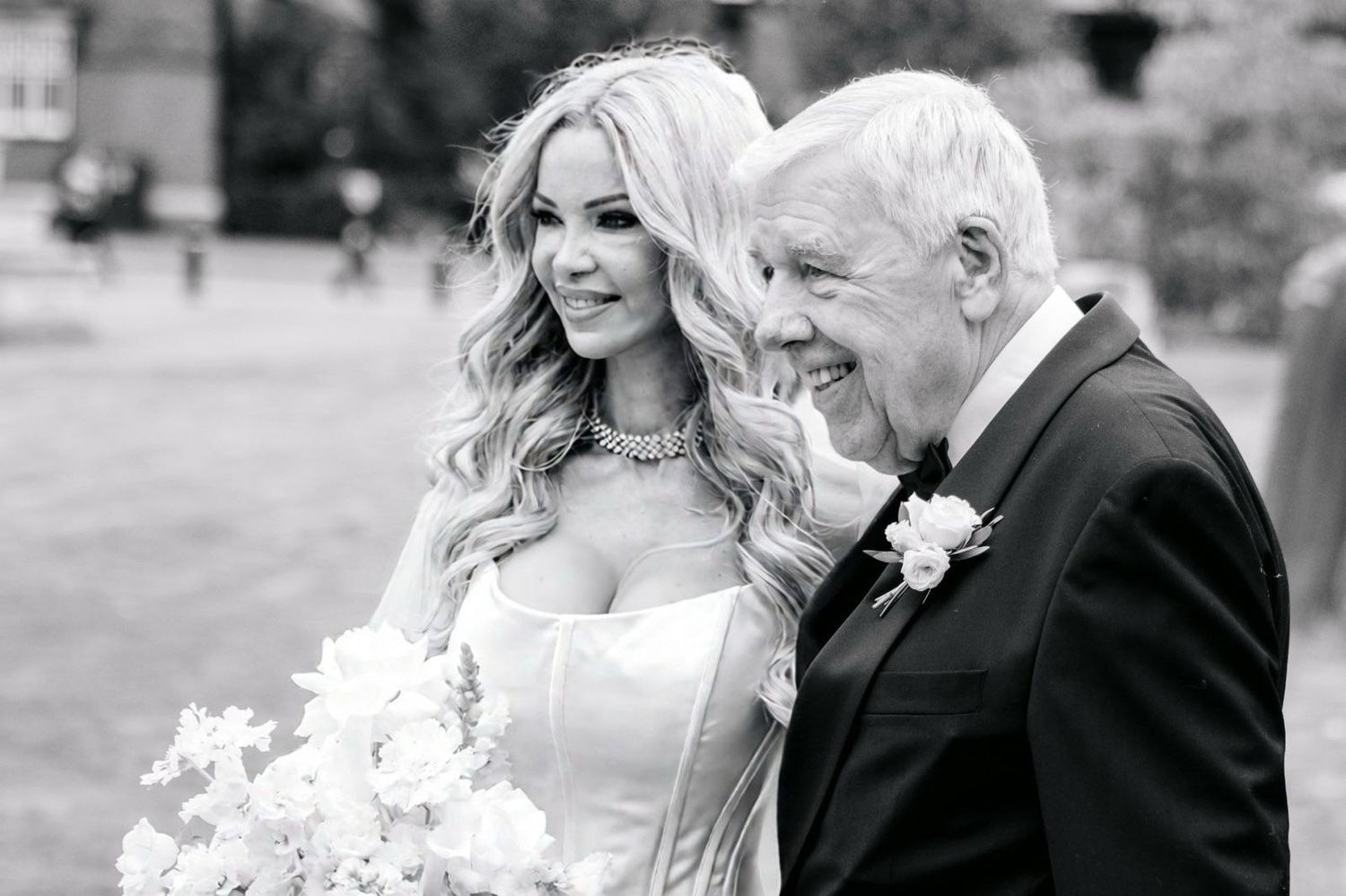 Black and white photo of a smiling bride in a strapless gown holding flowers, walking with an older gentleman in a tuxedo.