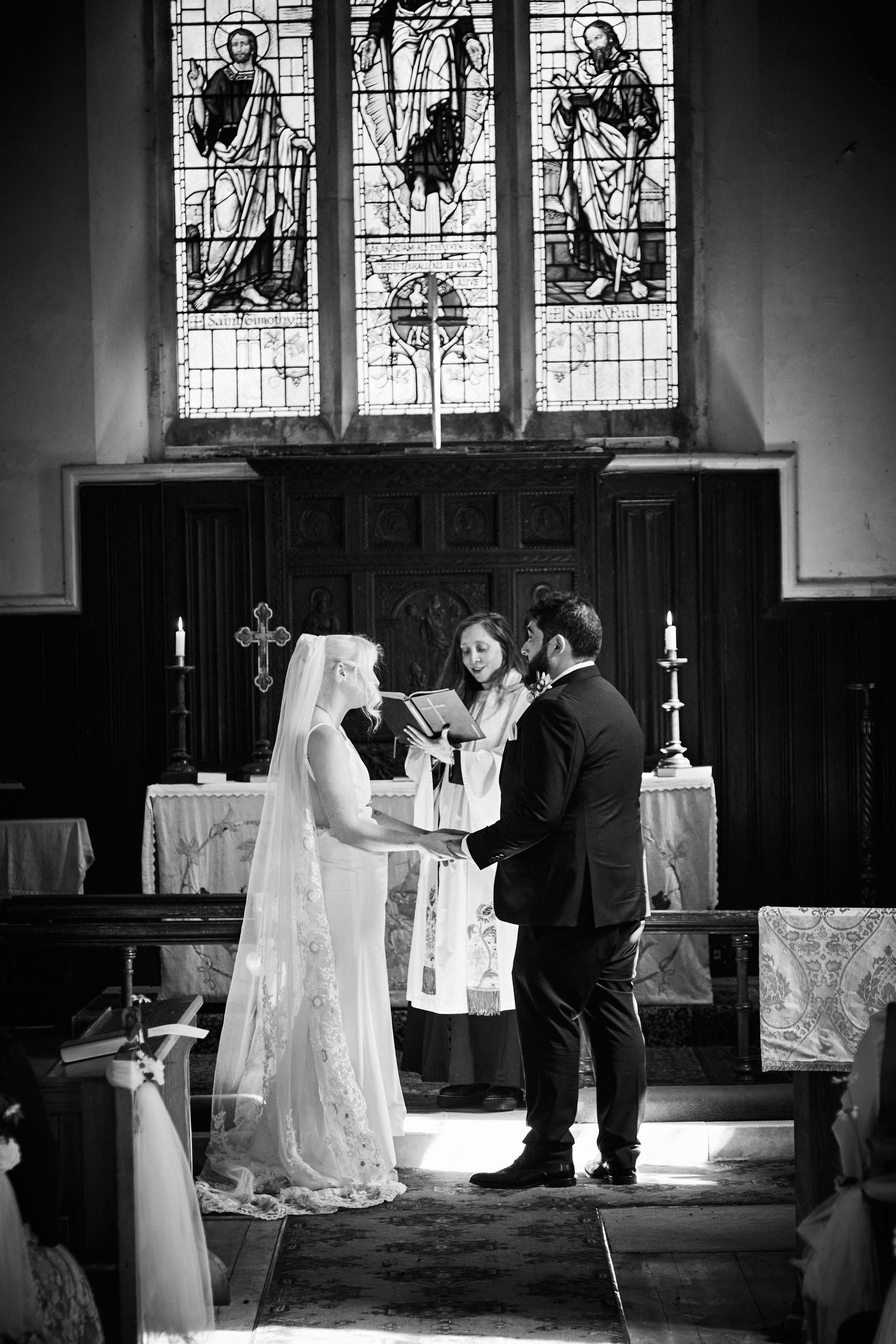 Black and white church wedding ceremony with couple exchanging vows before stained glass windows at altar.