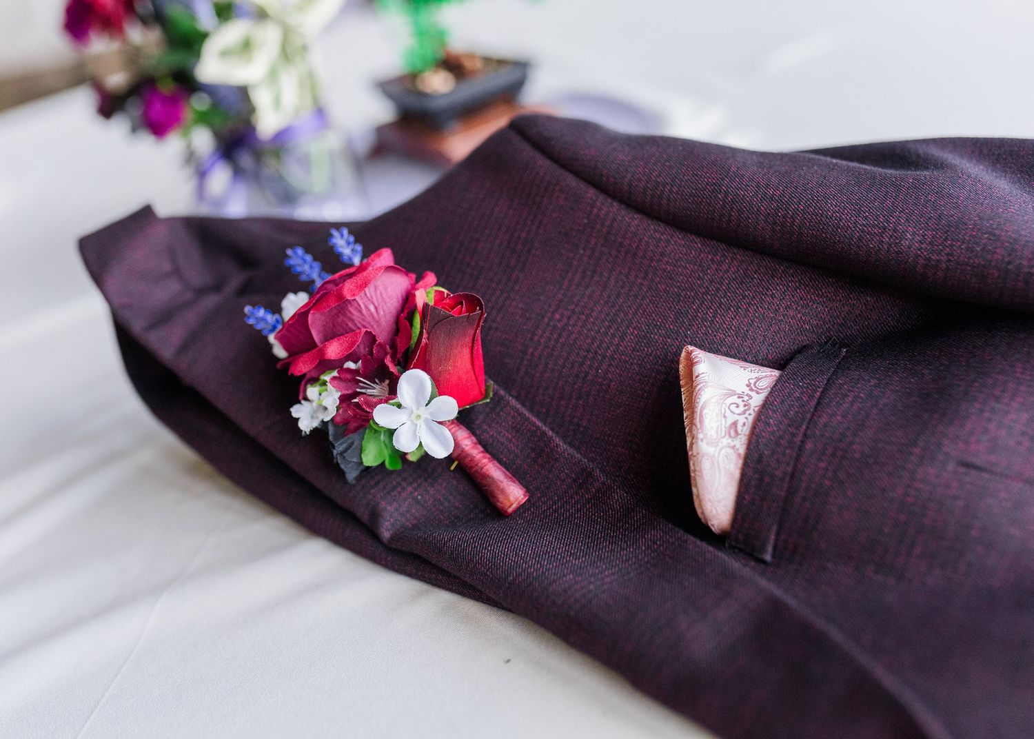 Dark plum suit jacket with red rose boutonniere featuring lavender and white flowers, displayed on a white surface.