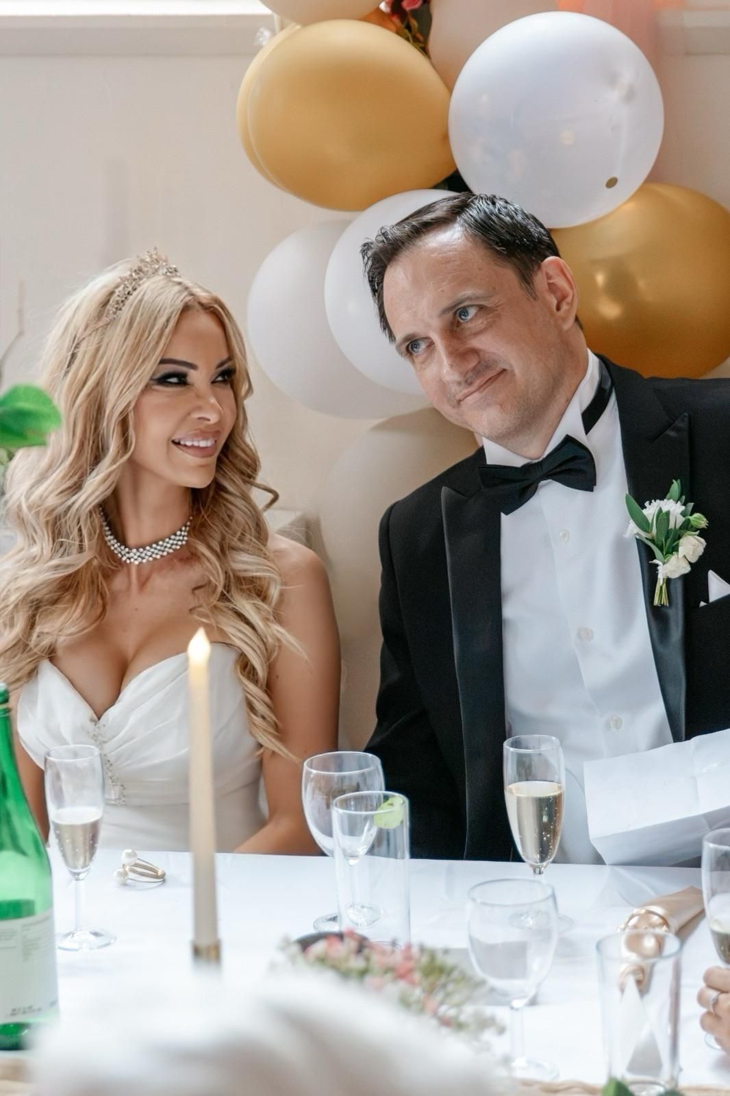 Bride and groom in formal attire sit at wedding reception table with gold and white balloons.