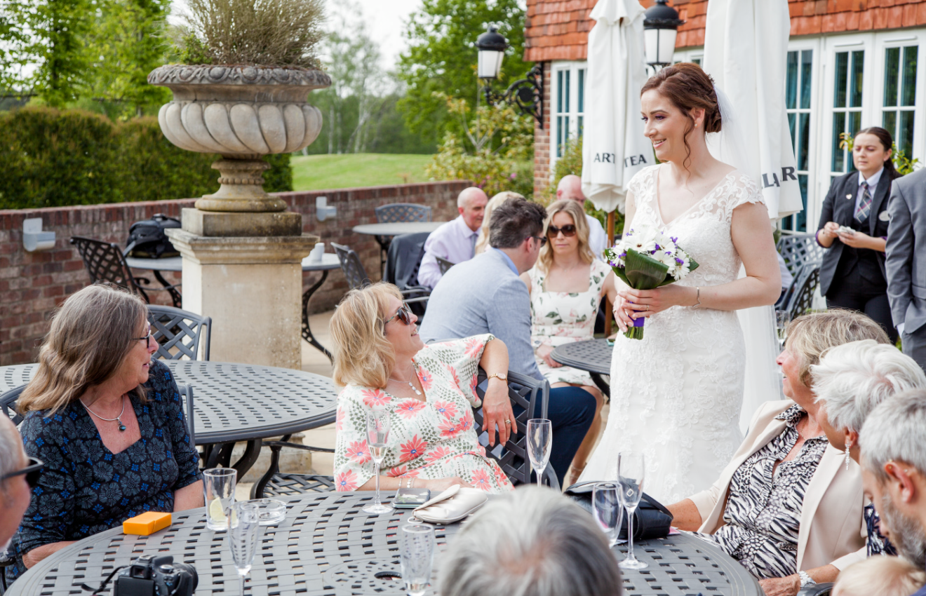 Bride in white gown mingling with seated wedding guests on sunny outdoor terrace during reception.