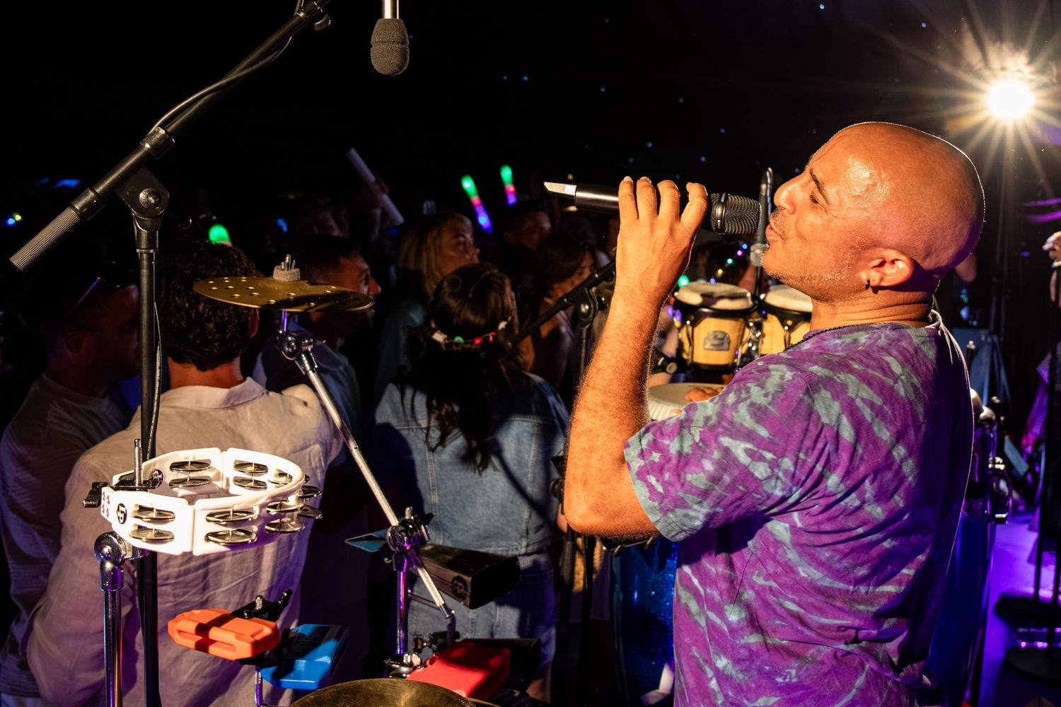 A bald musician in a purple tie-dye shirt plays a wind instrument energetically on a colorful lit stage.