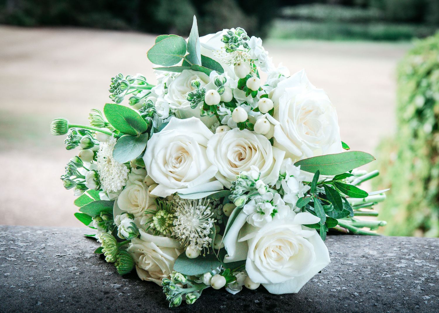 Elegant white rose bridal bouquet with greenery and small white flowers resting on a stone surface.