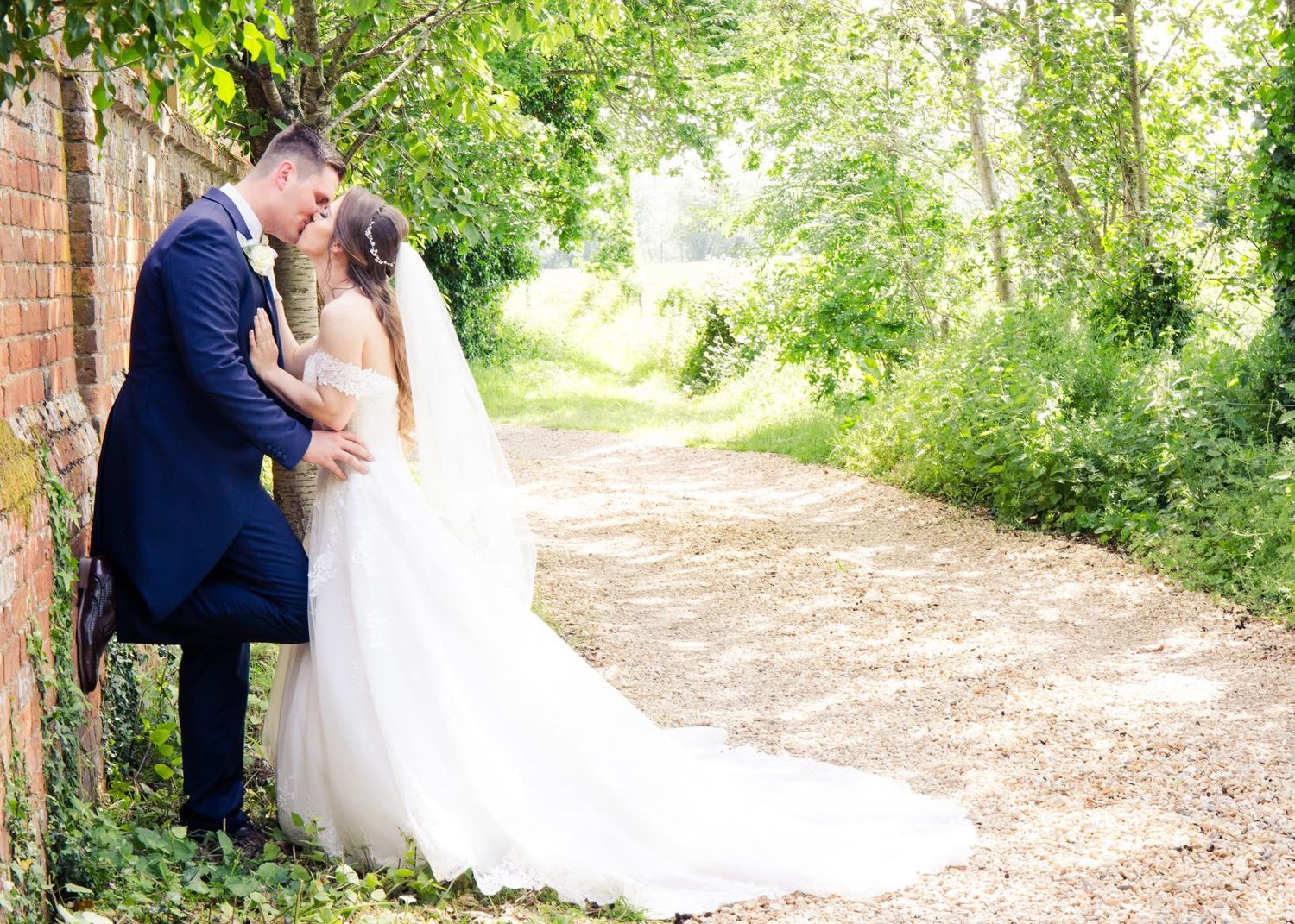 Bride and groom kissing beside brick wall on tree-lined path in wedding attire.