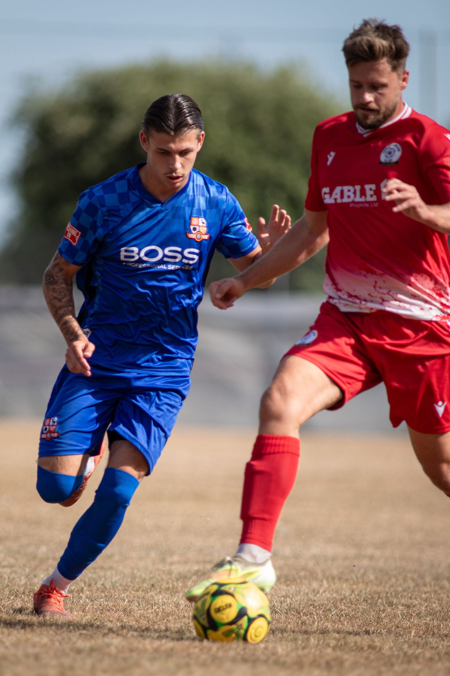 Two footballers in blue BOSS and red kits compete for the ball on a dry summer pitch.