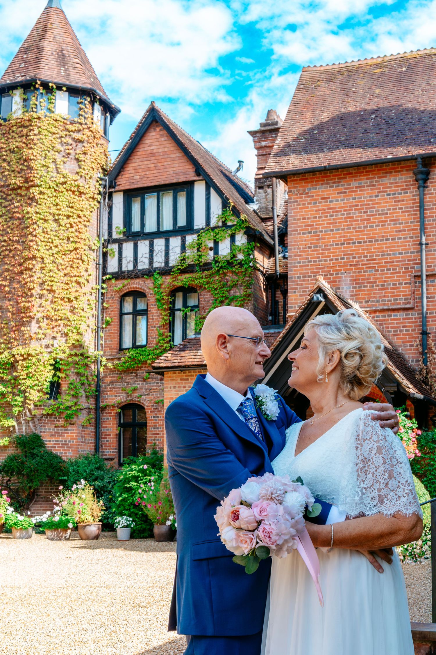 Bride and groom embrace in front of a charming brick Tudor manor with ivy-covered tower on their wedding day.